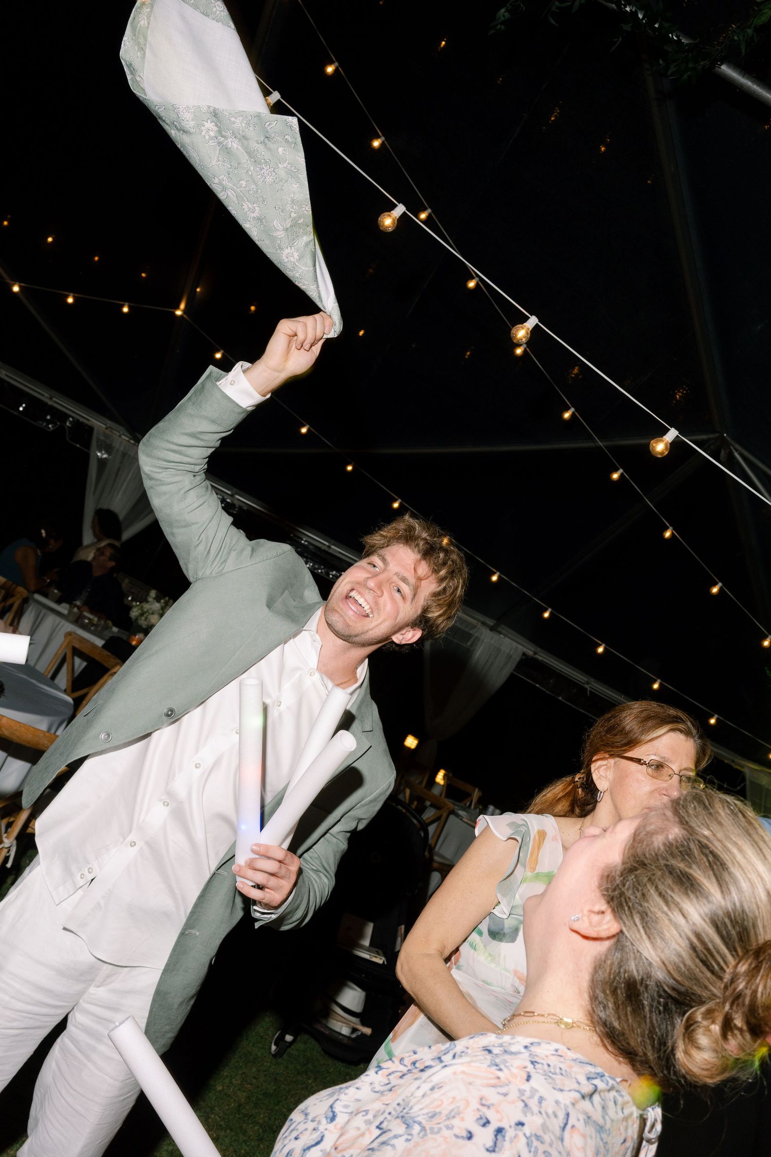 Groom in sage green suit enthusiastically waving napkin while dancing with guests under string lights at wedding reception.