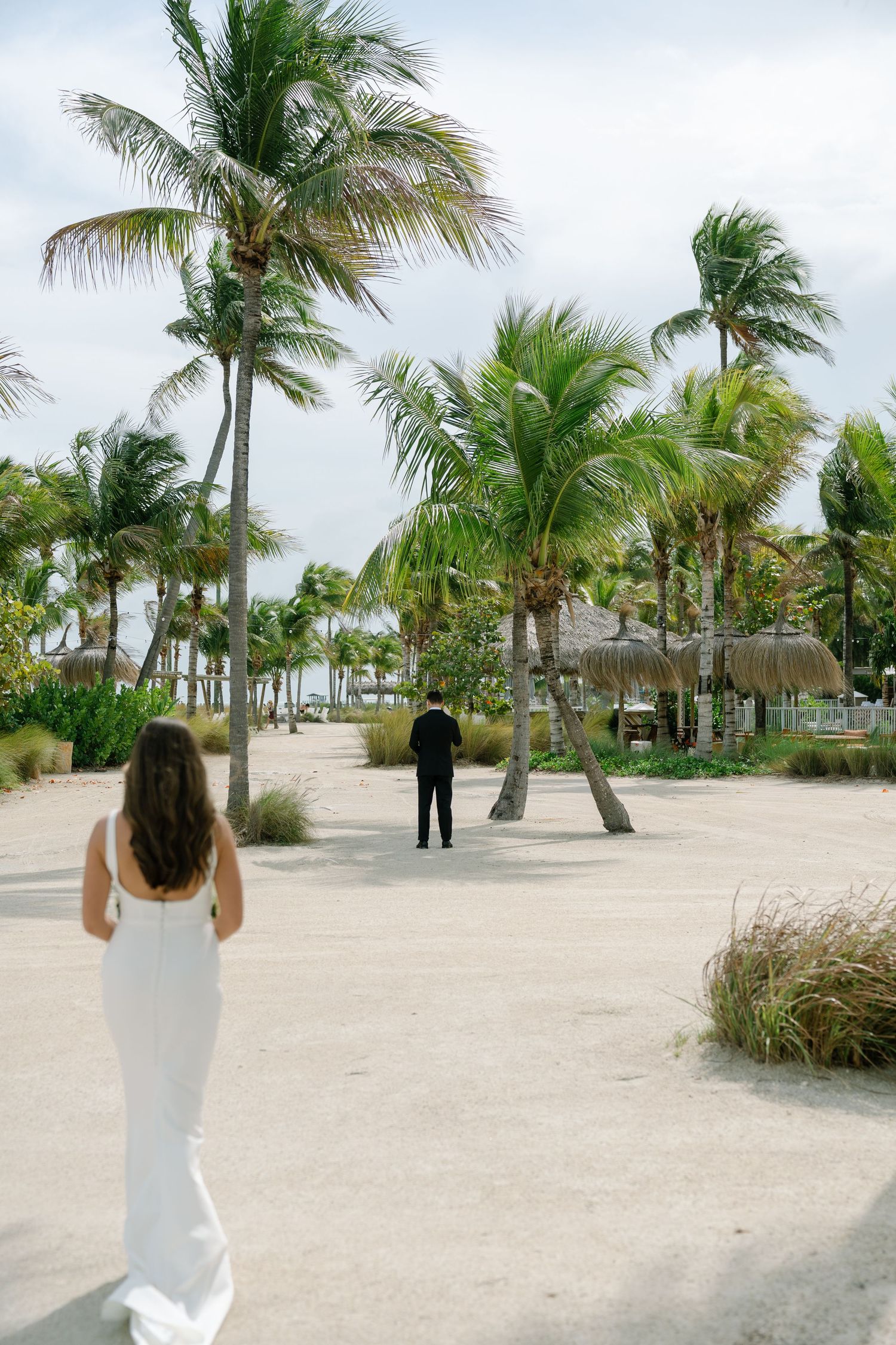 Bride in white gown walking toward groom on sandy beach path lined with palm trees.
