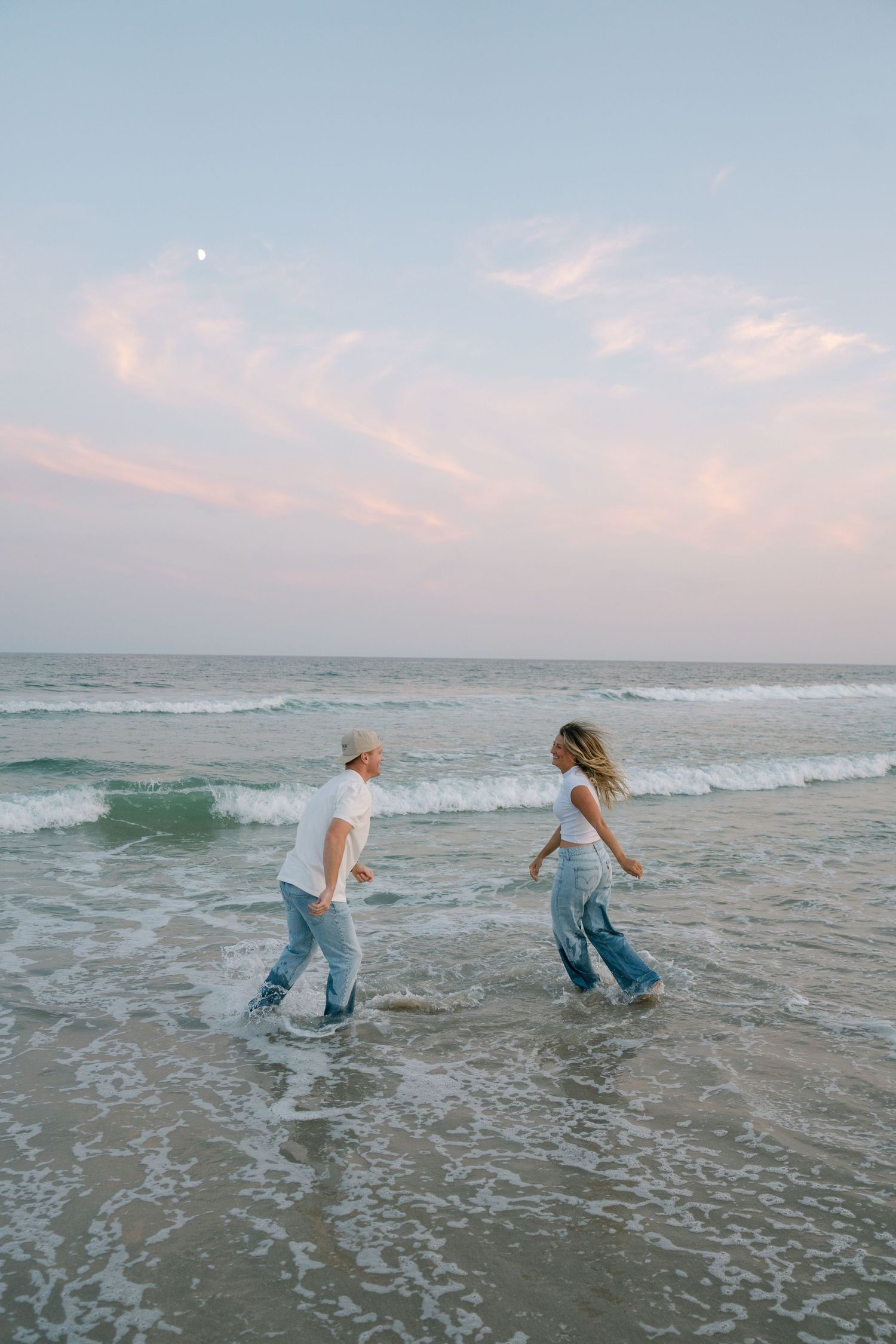 Two people in jeans splash and play together in ocean waves at sunset on a peaceful beach.