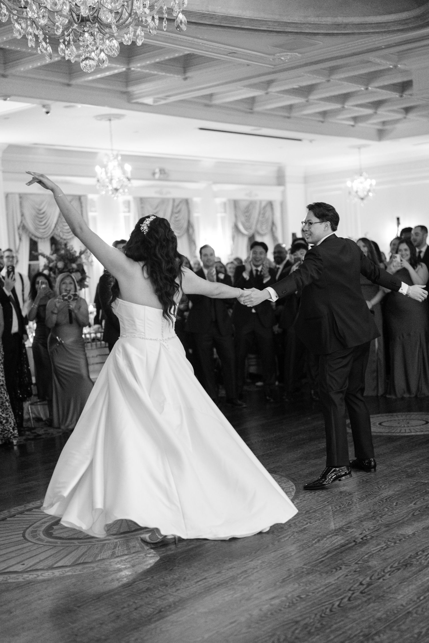Couple dancing in elegant ballroom with spinning white dress and chandelier overhead.