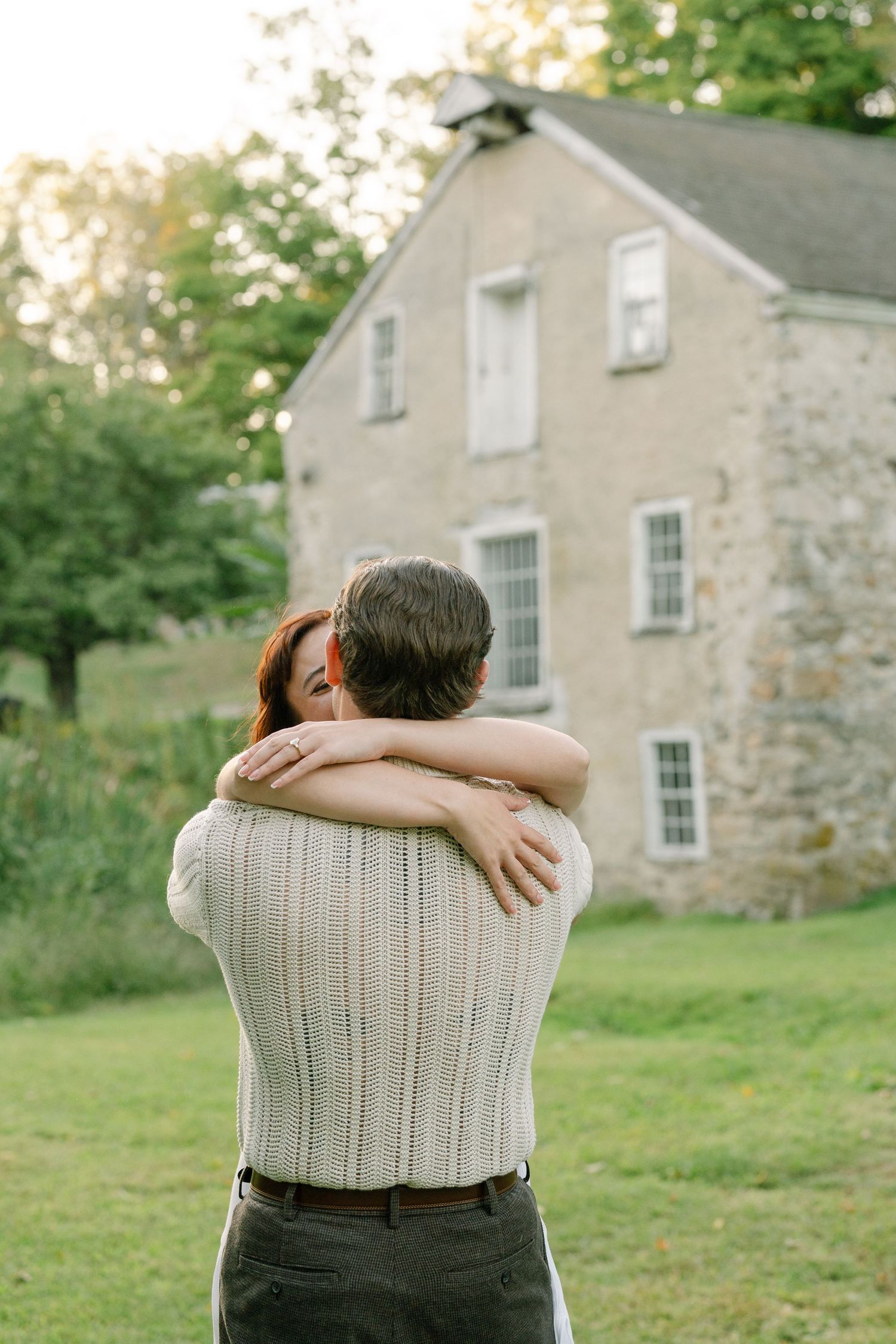 A couple embraces tenderly on a green lawn in front of a historic stone building during golden hour.