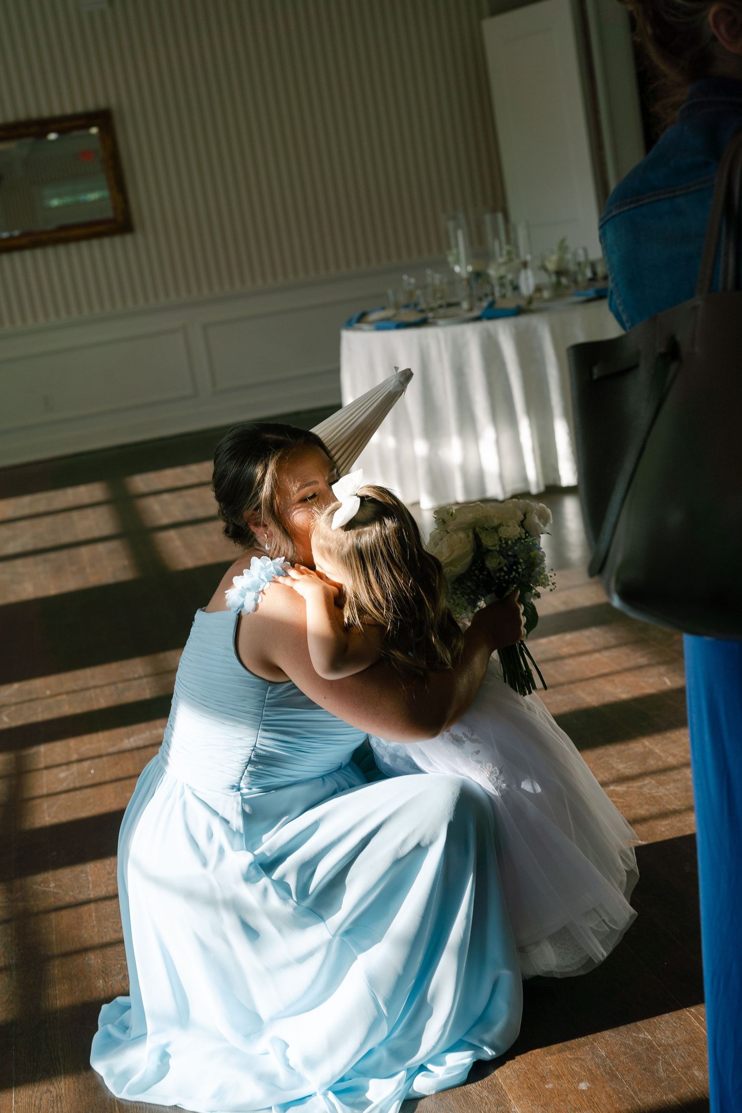 Bridesmaid in light blue gown kneels to hug flower girl wearing unicorn hat inside wedding reception venue.