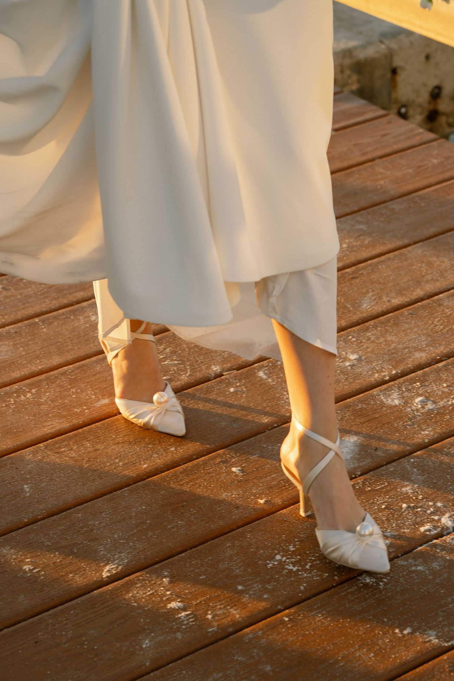Detail shots of bridal shoes and dress hem on wooden dock at sunset.