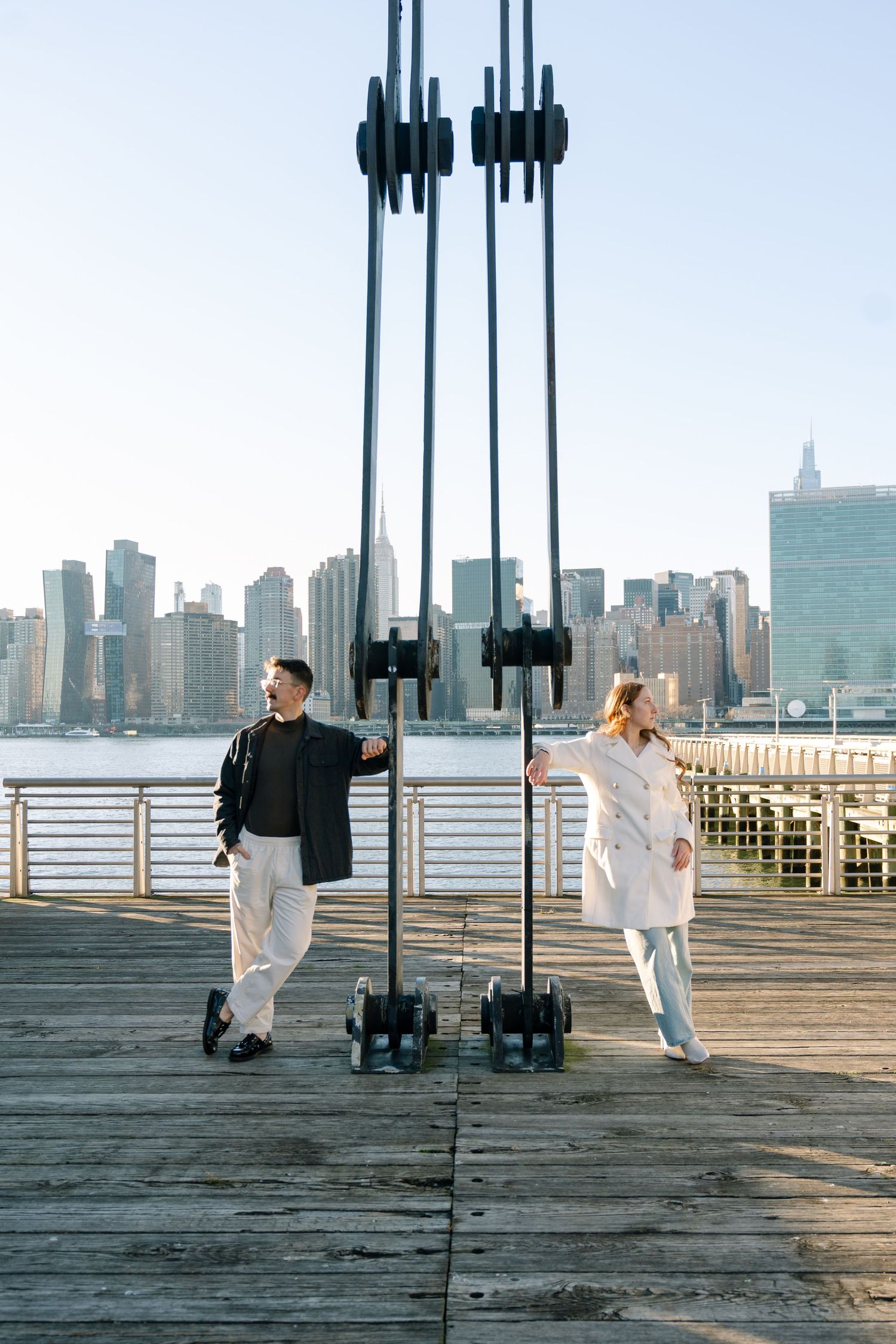 People in white clothing walk along a wooden pier with the New York City skyline visible in the background.