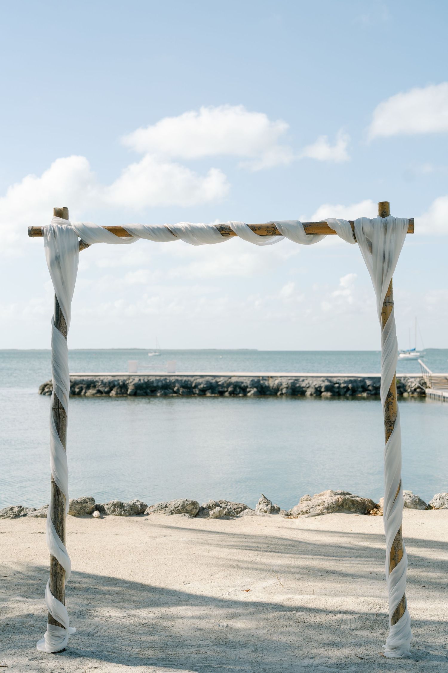 A white and striped wooden arch structure on a beach with a body of water in the background.