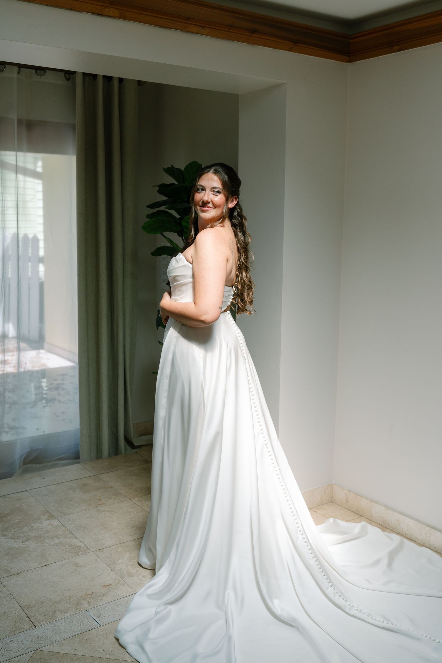 A woman in a white wedding dress stands in a room with a large window.