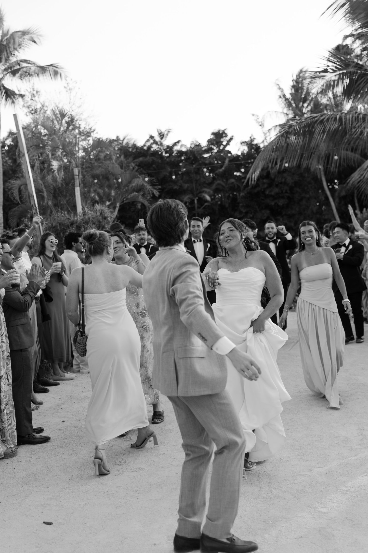 Black and white photo of bride and groom dancing outdoors at a tropical beach wedding with guests cheering.