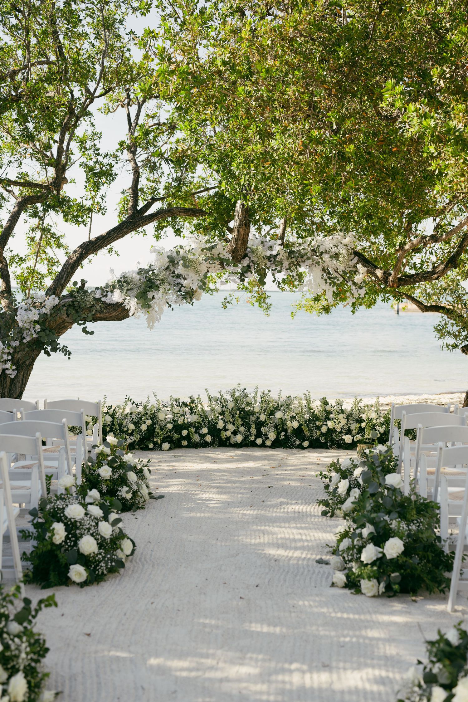 Elegant outdoor wedding ceremony setup with white chairs and lush floral aisle arrangements under a tree by the ocean.