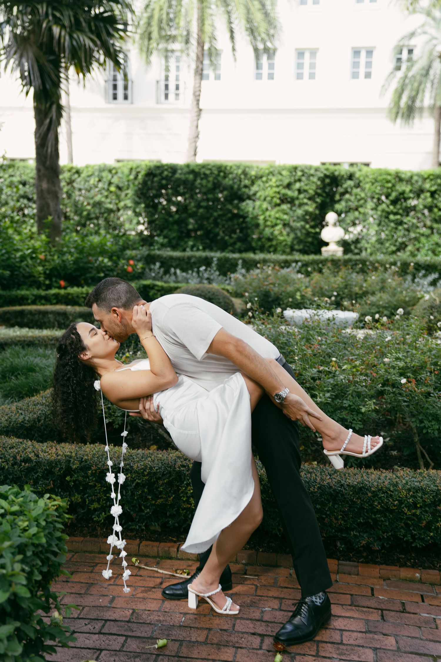 A romantic dip pose on a brick path surrounded by manicured gardens and palm trees.