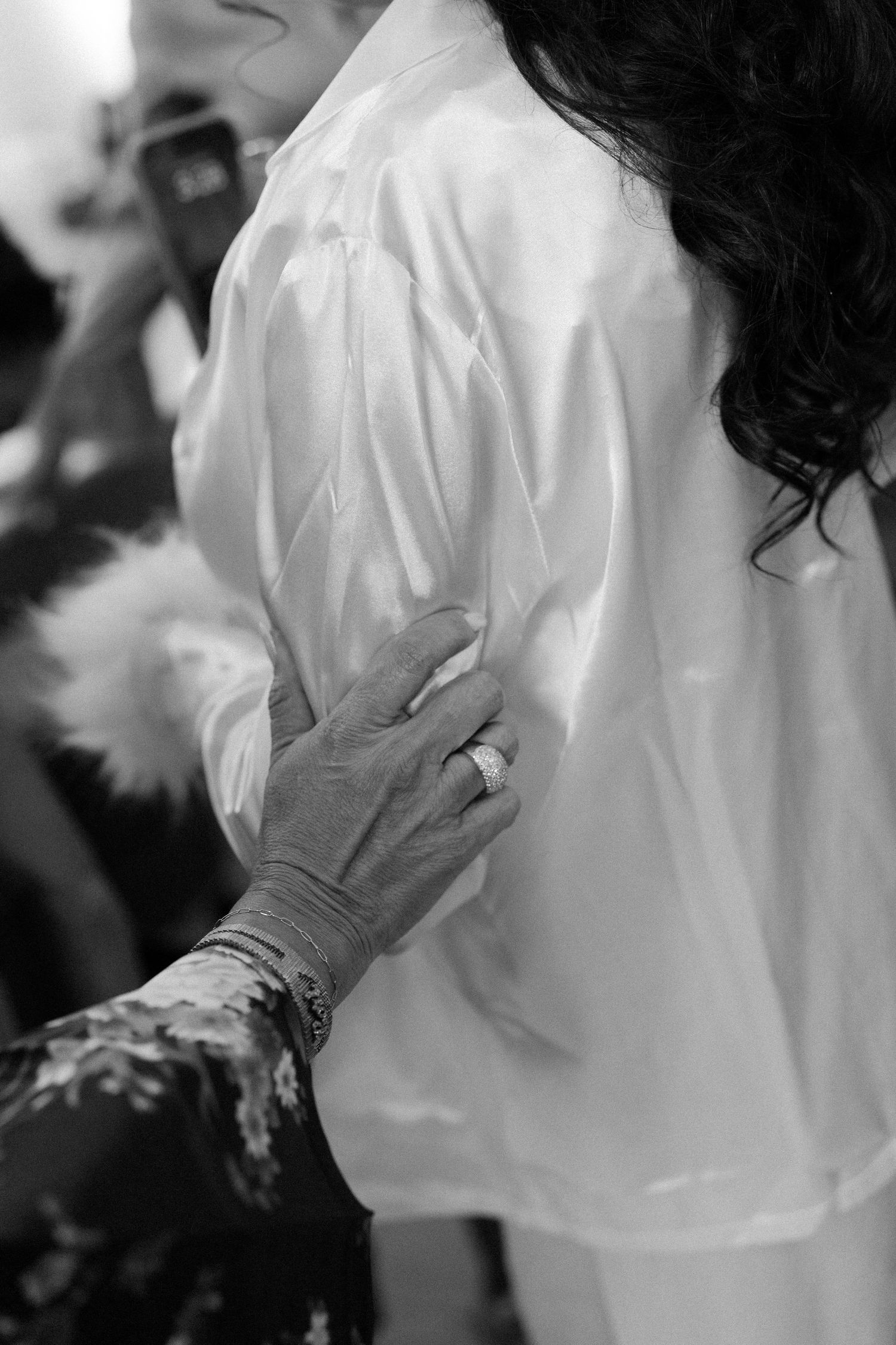 A black and white closeup of a hand with tattoos resting on a white fabric-covered shoulder.