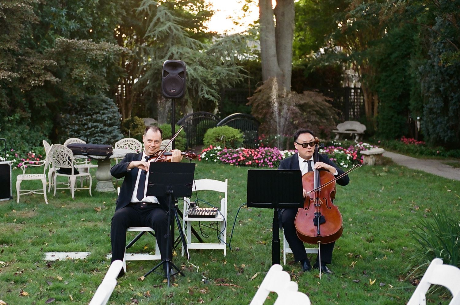 Musicians perform outdoors at a garden wedding ceremony with white chairs and natural greenery surroundings.