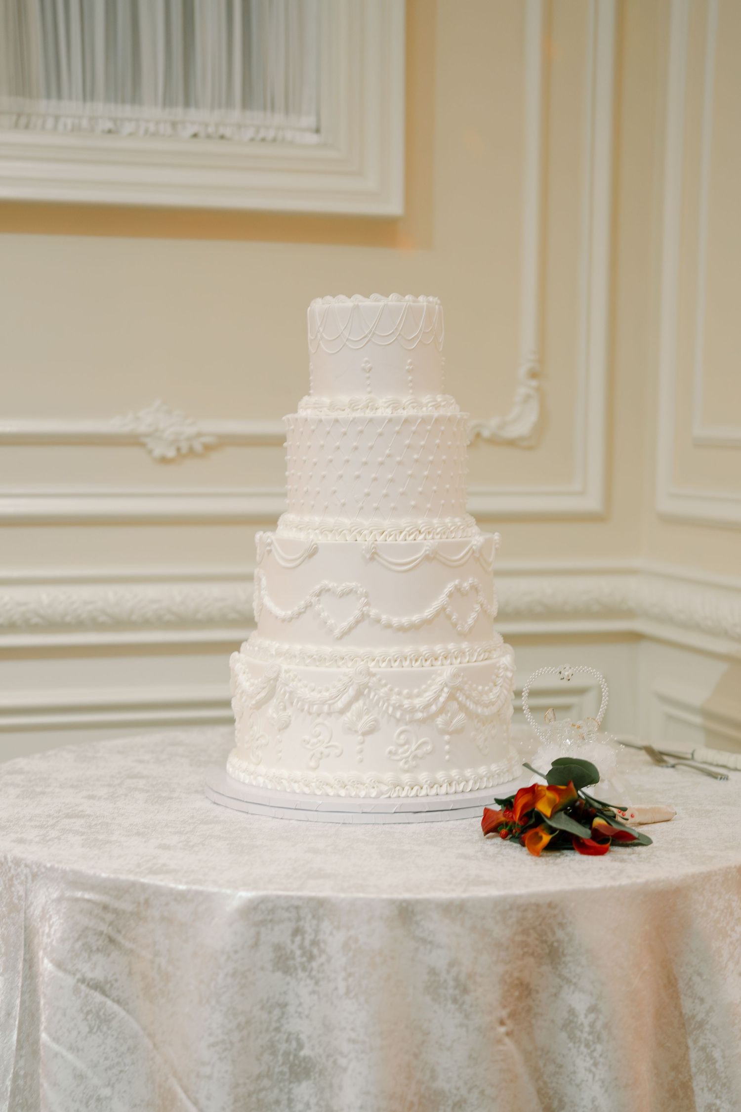 An elegant white four-tier wedding cake with lace detailing sits on a silver tablecloth.