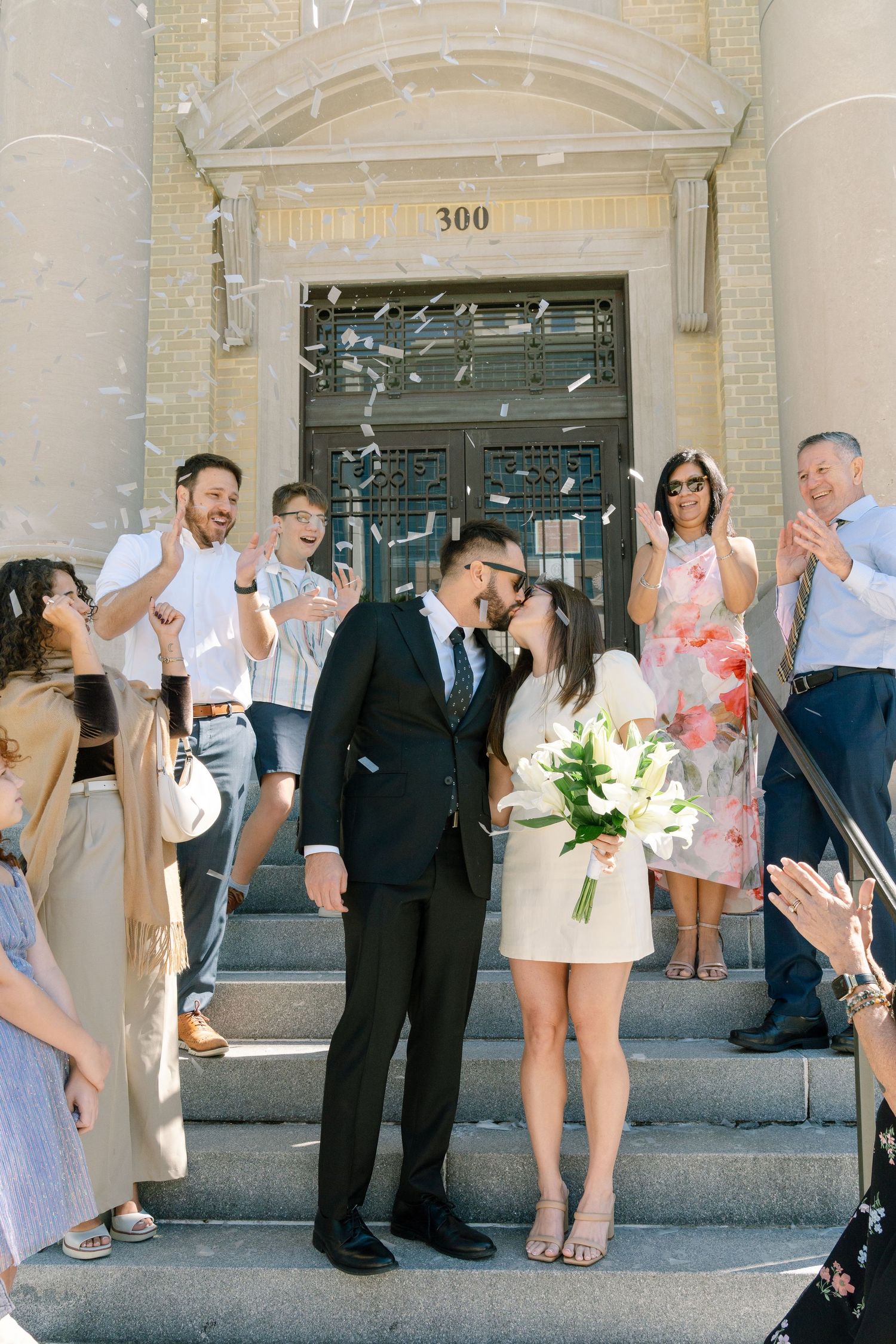 Wedding party members celebrate with joyful poses and gestures on the steps of a grand stone building.