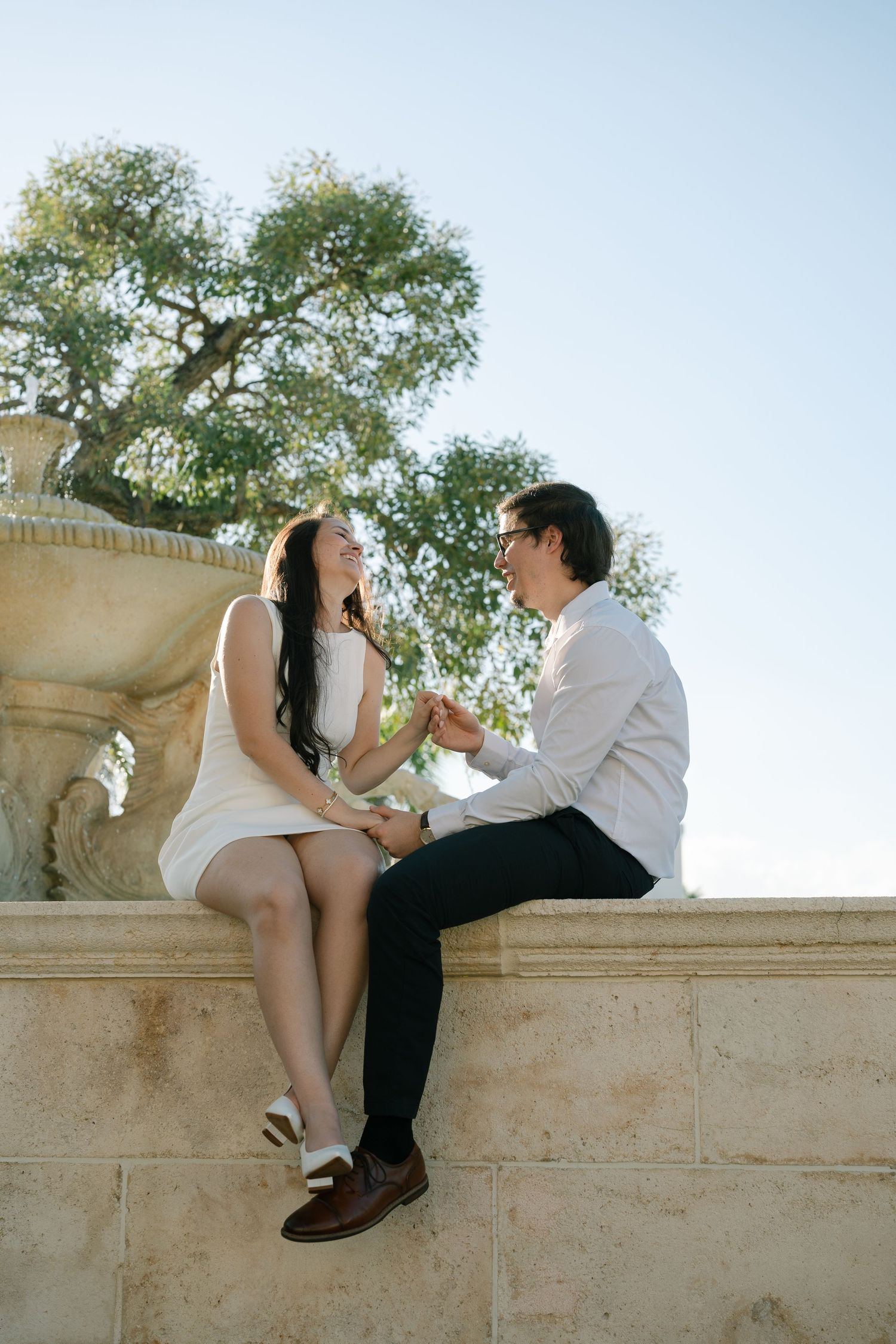 A romantic couple sits together on a stone ledge under a tree on a sunny afternoon.