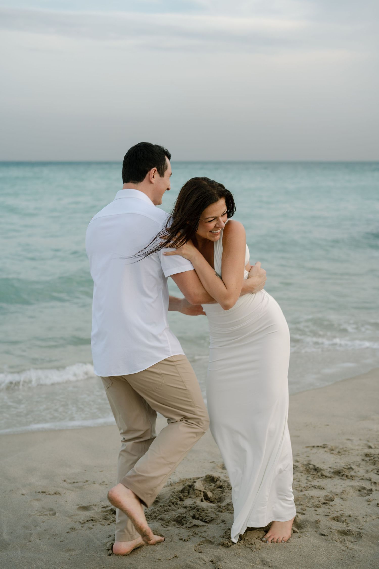 A playful beach photoshoot with couple in white dress and casual attire on sandy shore.