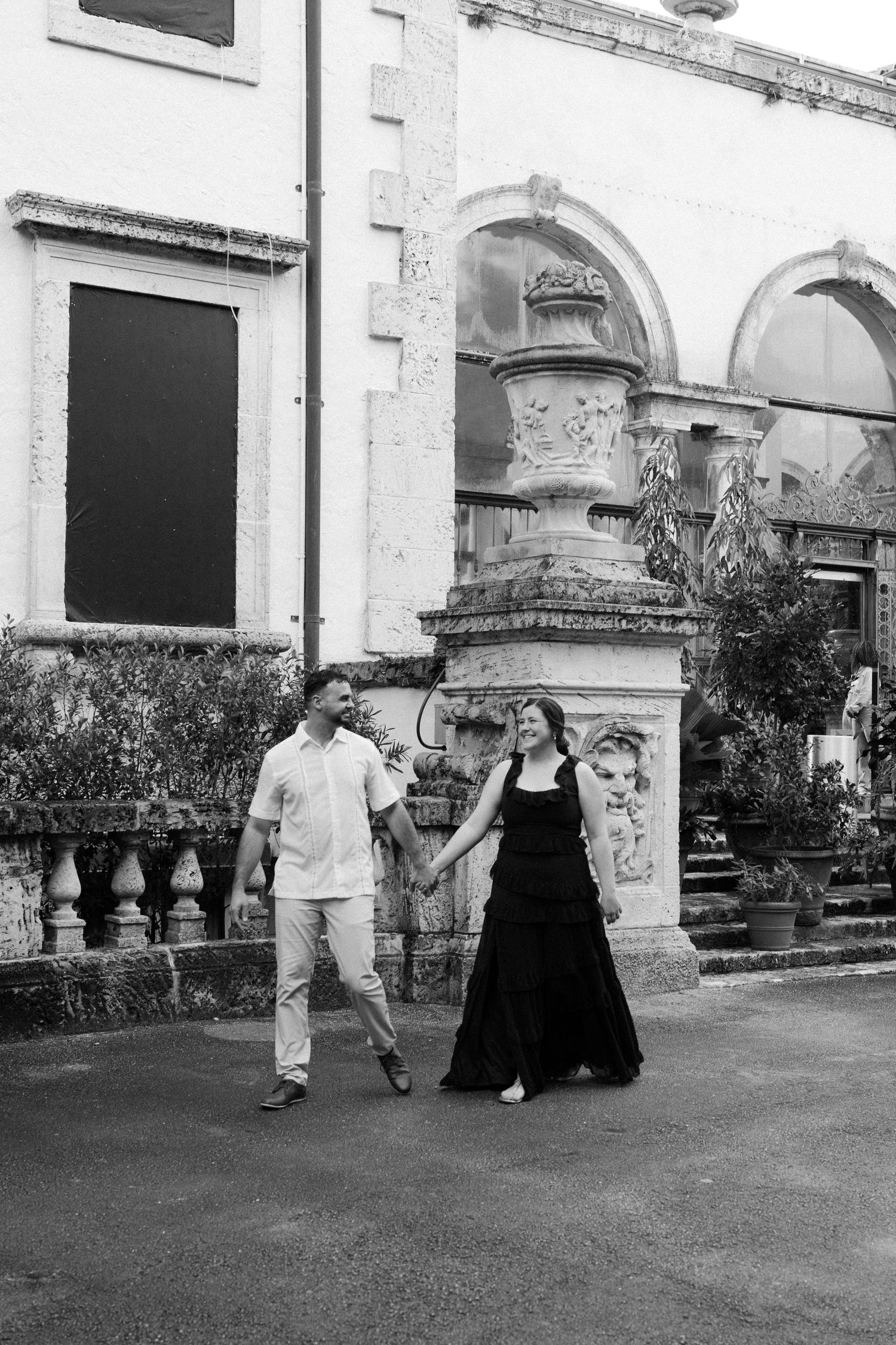 A couple walks through a grand courtyard with ornate stone architecture and arches in black and white.
