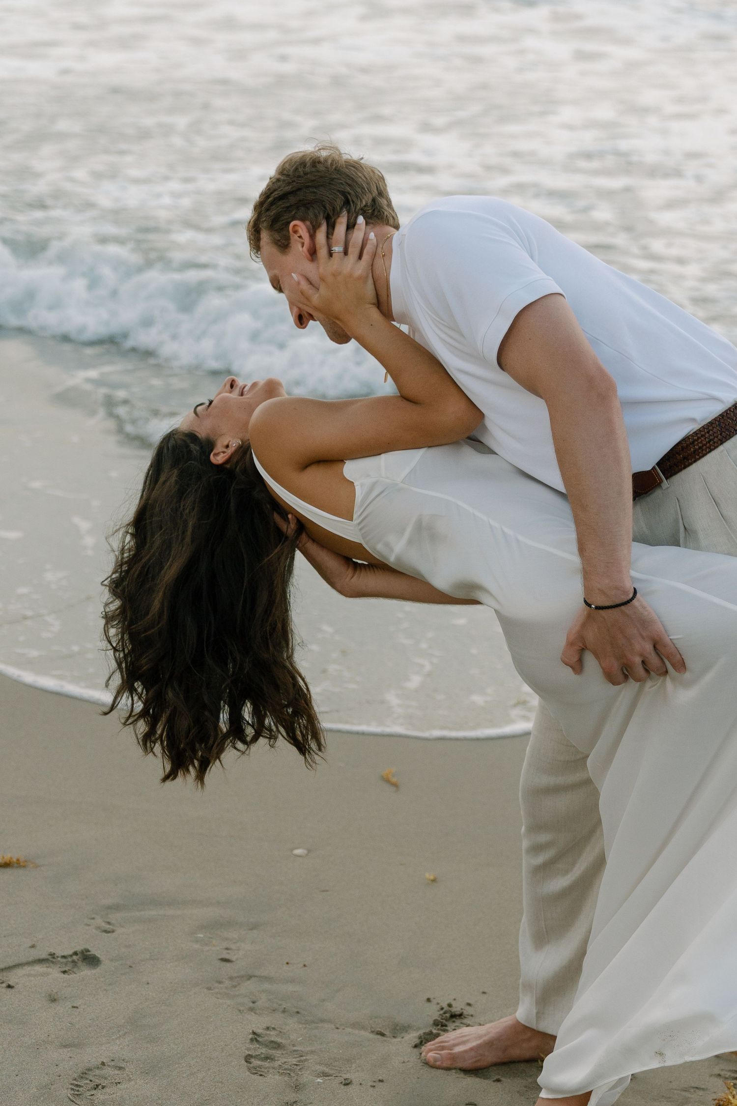 Couple sharing a romantic dip kiss on a sandy beach with ocean waves, woman in white dress, man in white shirt.