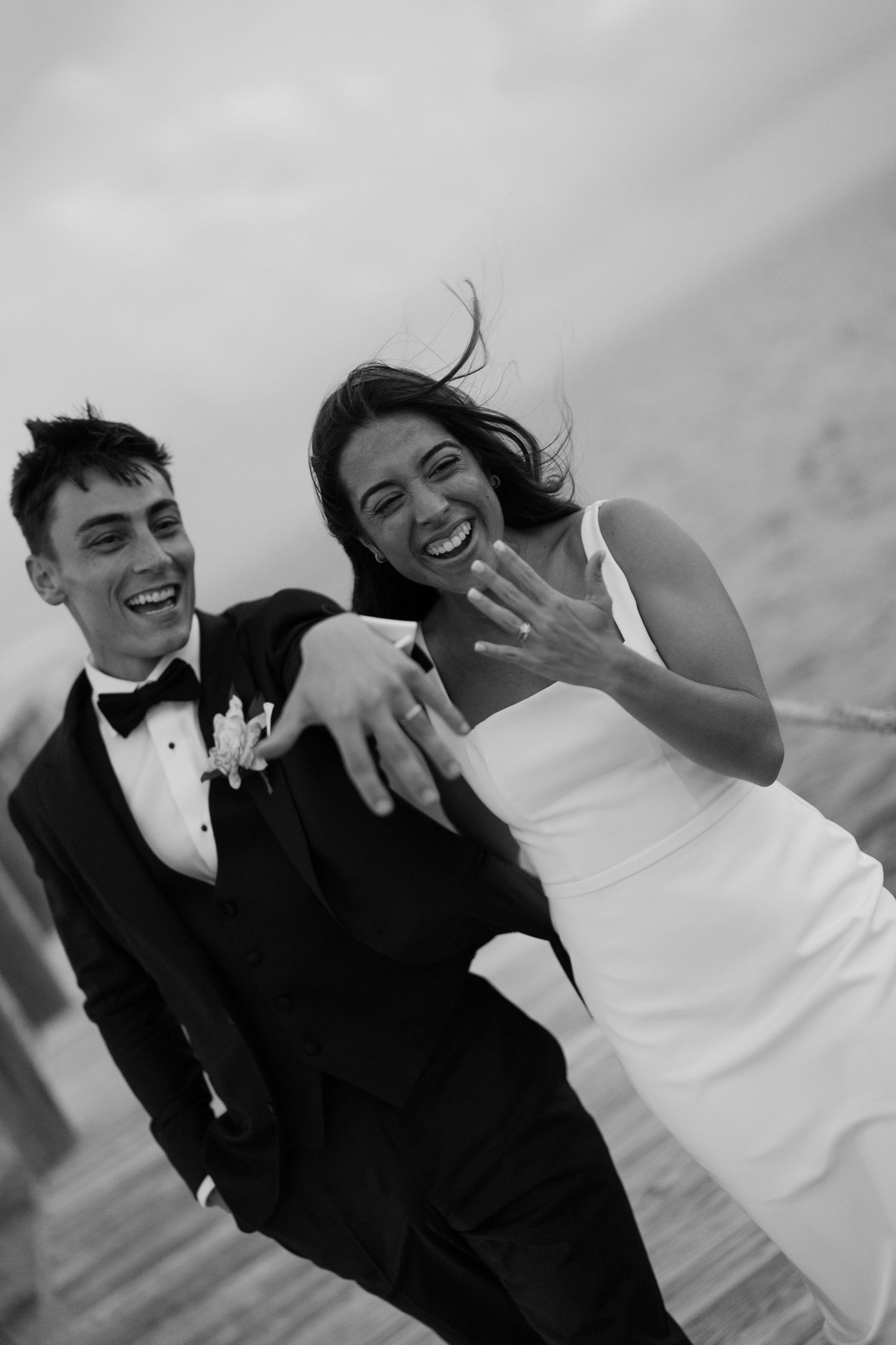 Joyful newlyweds in black and white, bride shows off wedding ring while laughing with groom on windy waterfront.