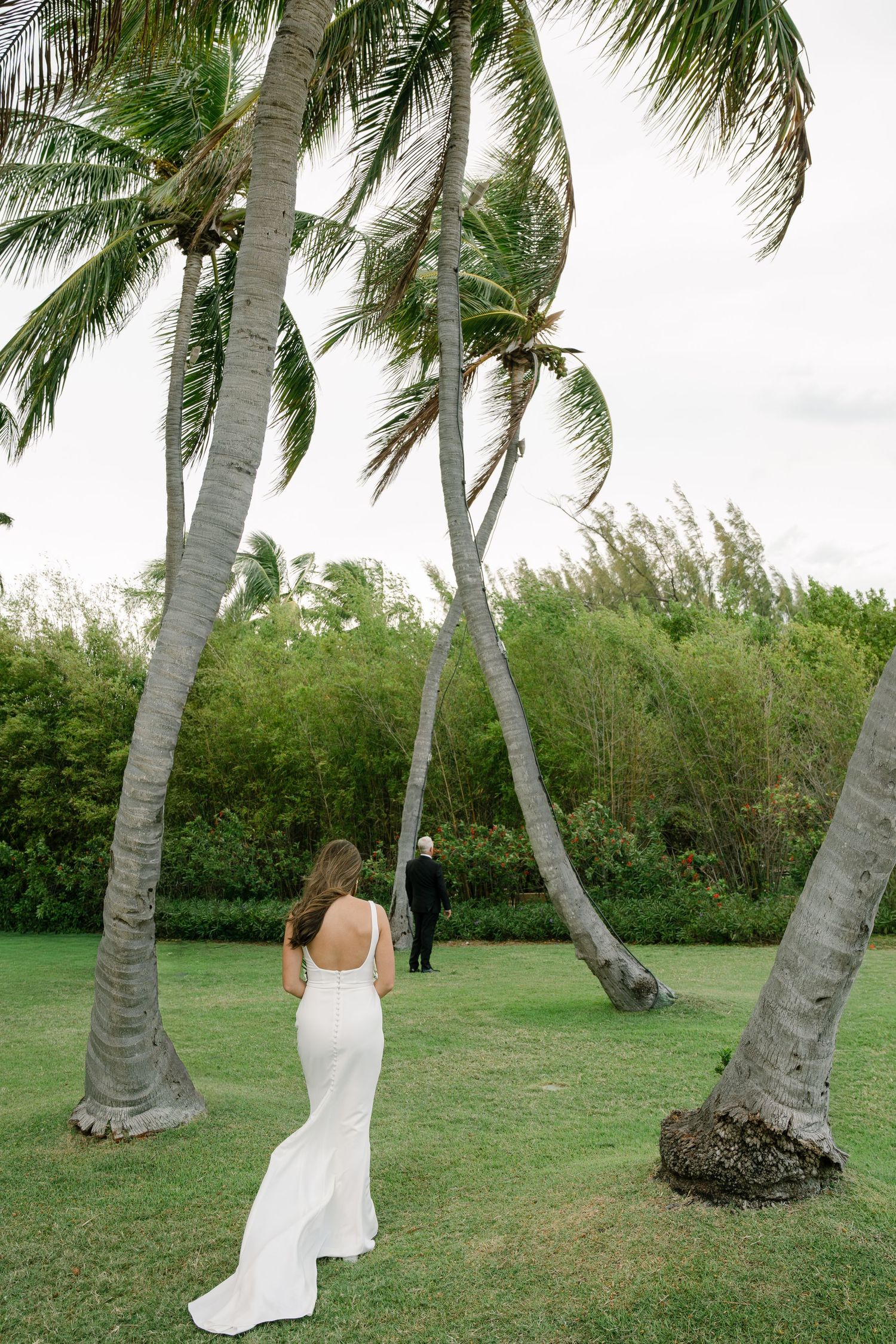Bride in backless white gown walks toward groom between leaning palm trees at tropical outdoor wedding venue.