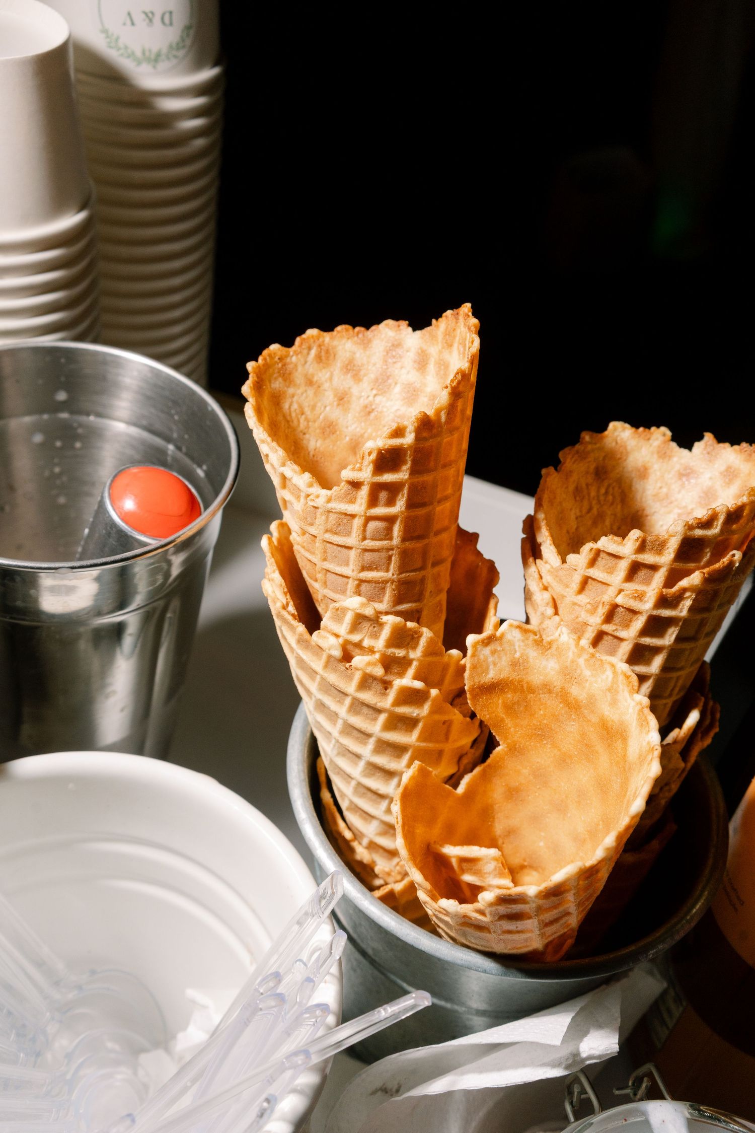 Empty waffle ice cream cones in a metal holder beside stacked cups and a silver bucket at a dessert station.
