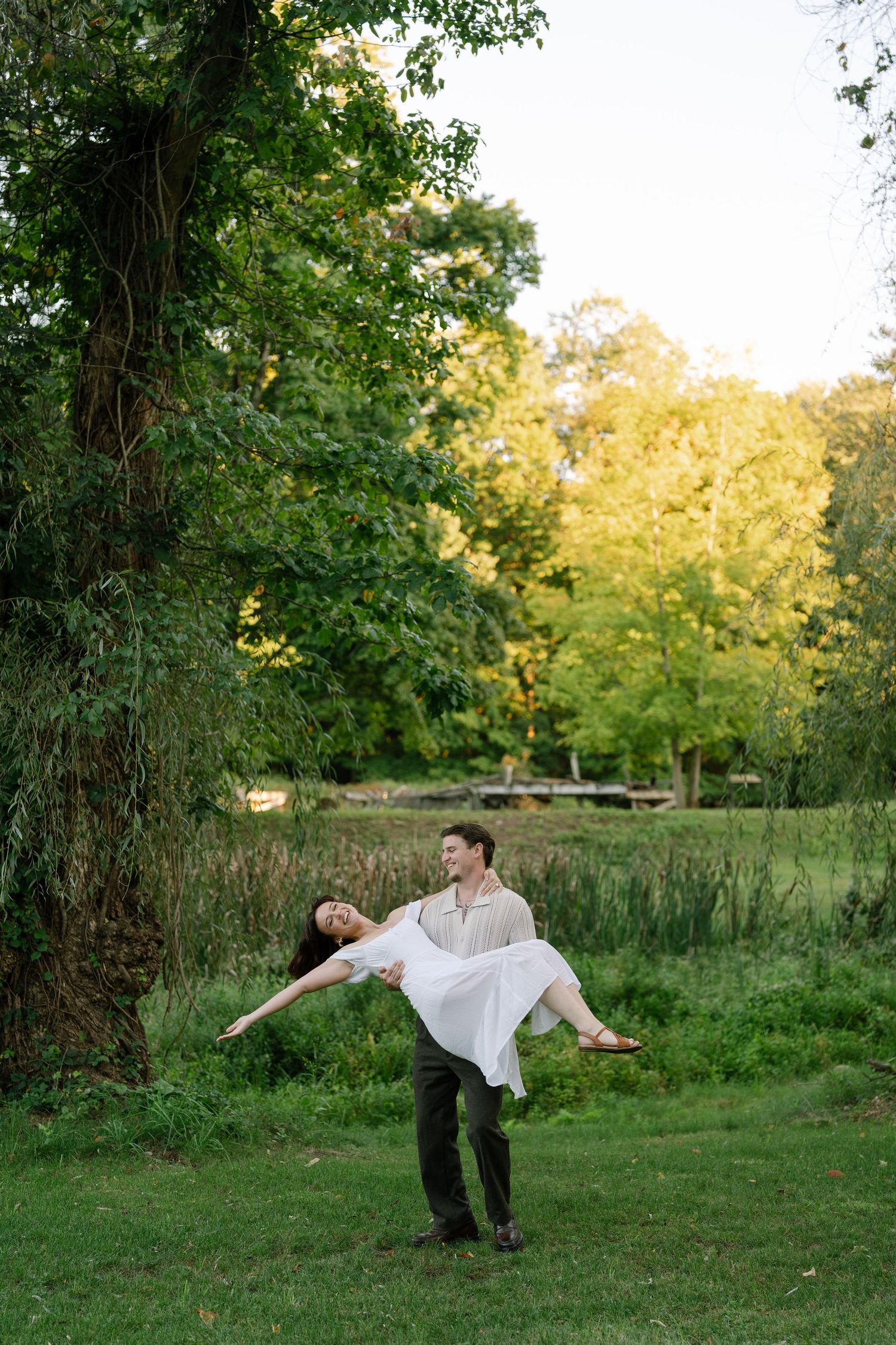 A man joyfully spins a woman in a white dress in a lush green park during golden hour.