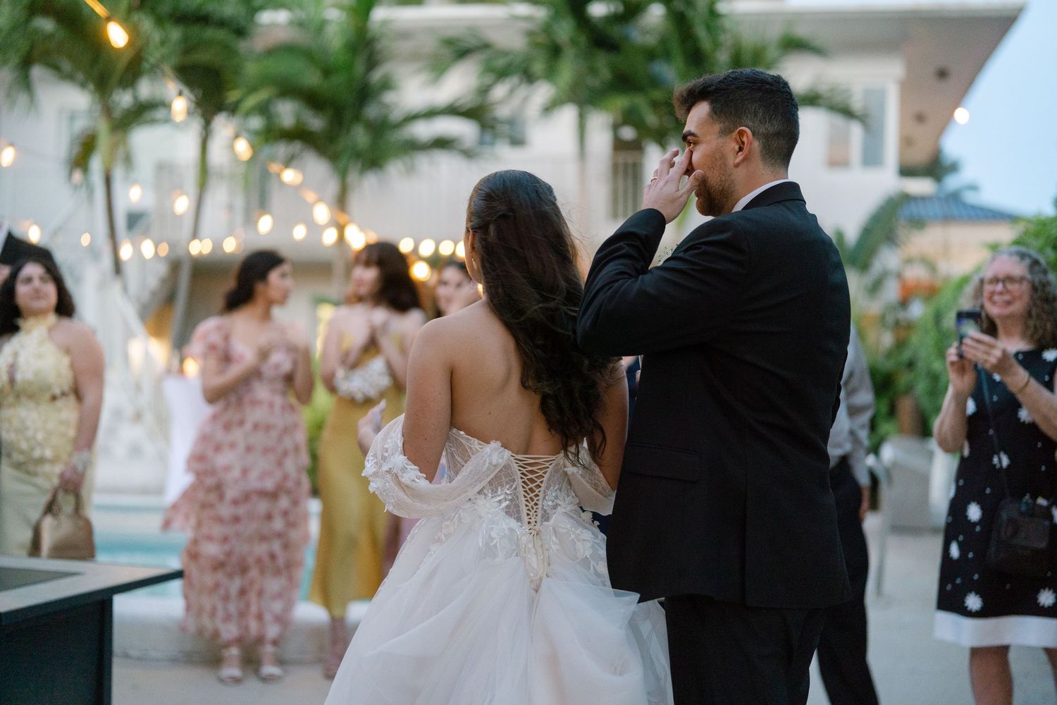 Bride and groom share emotional first dance outdoors at elegant wedding reception with string lights and guests.