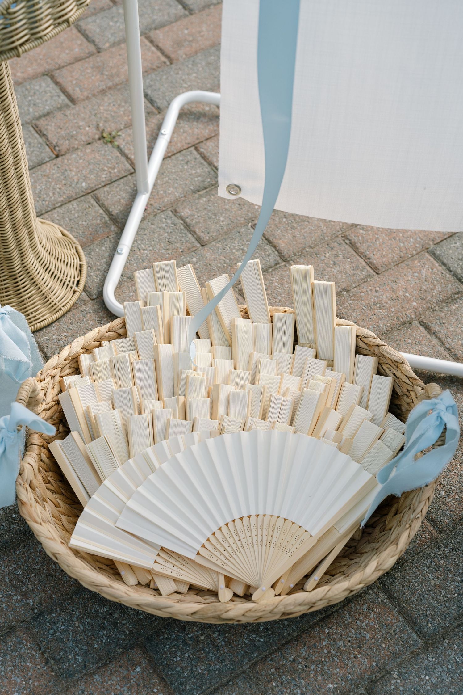 A wicker basket filled with white folding hand fans displayed outdoors at a wedding event on brick pavement.