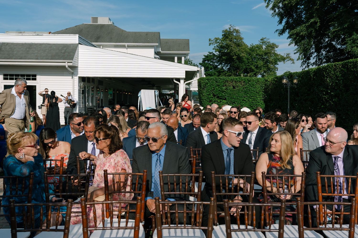 Guests seated in wooden chairs at an elegant outdoor wedding ceremony with lush green hedges.