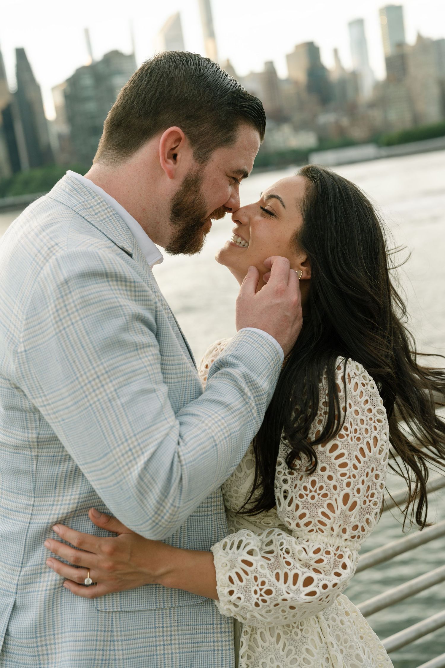 Couple nearly kissing with NYC skyline backdrop, man in plaid blazer tenderly holding woman's face near the waterfront.