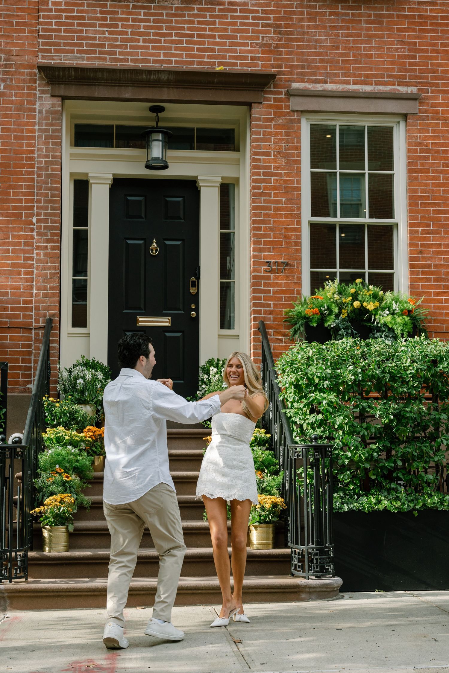 Couple dancing and laughing together outside a charming brick townhouse with a black door and lush greenery.