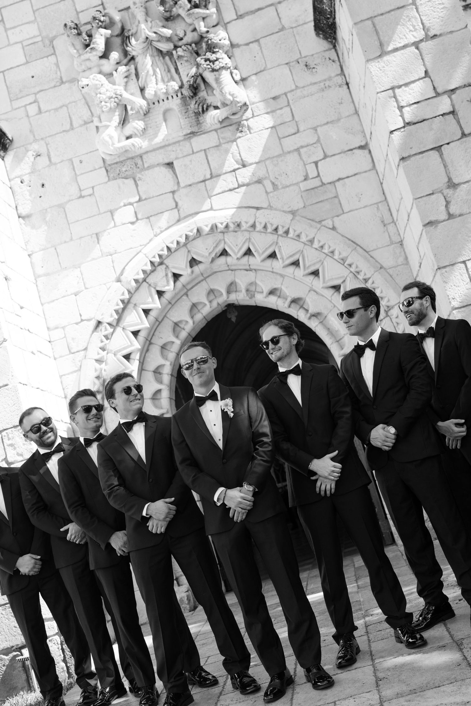 Black and white photo of groom and groomsmen in tuxedos and sunglasses posing outside stone church entrance.