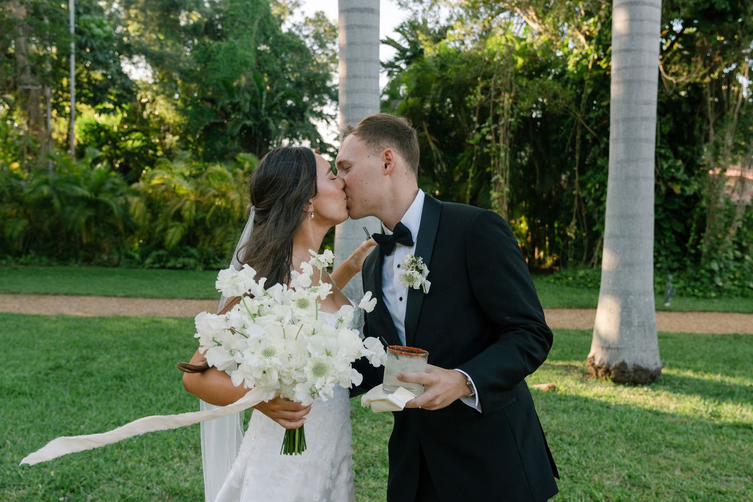 Bride and groom share romantic kiss holding white orchid bouquet in tropical garden.