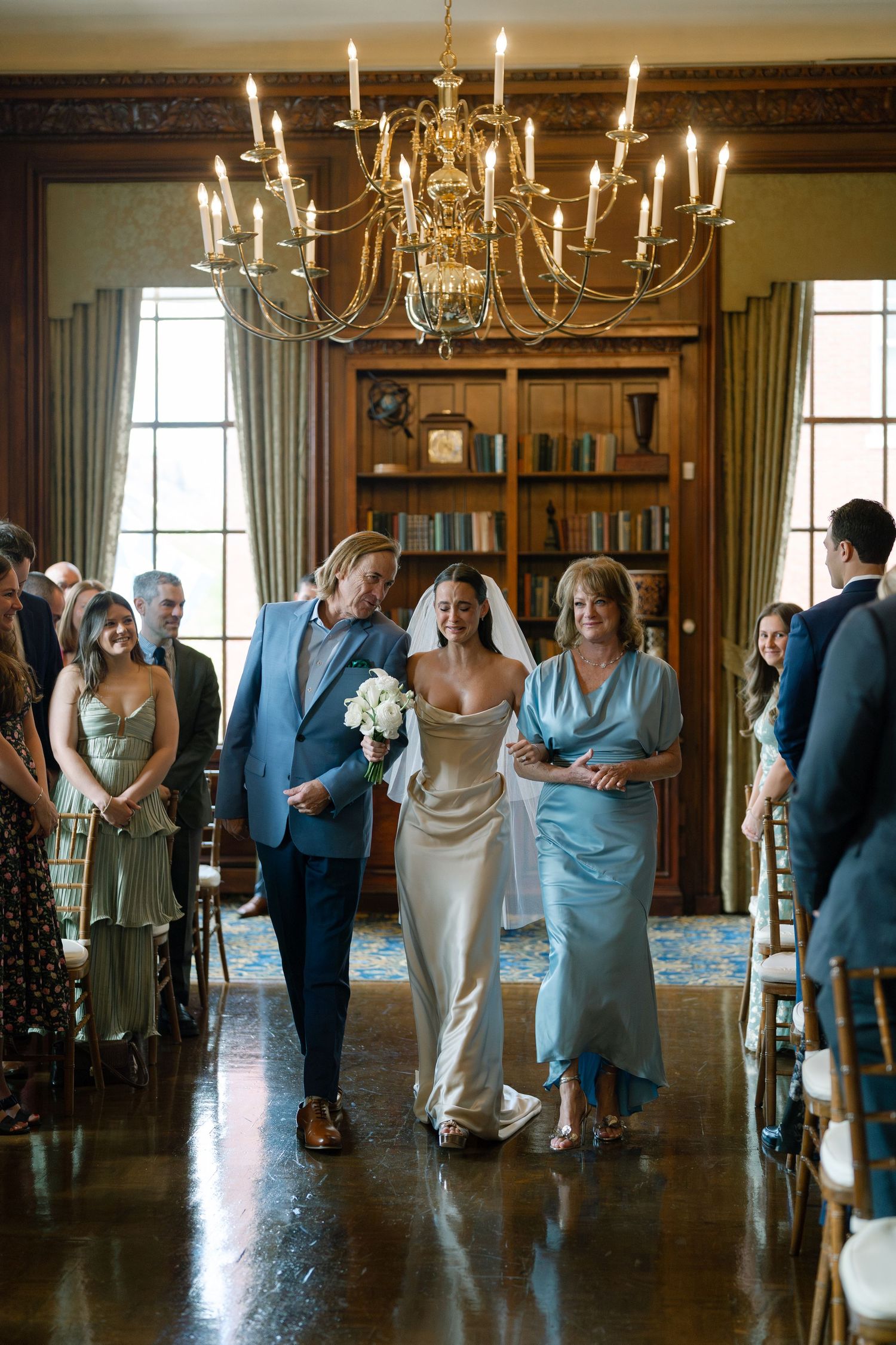 Bride walks down the aisle escorted by parents in elegant wood-paneled venue with gold chandelier.