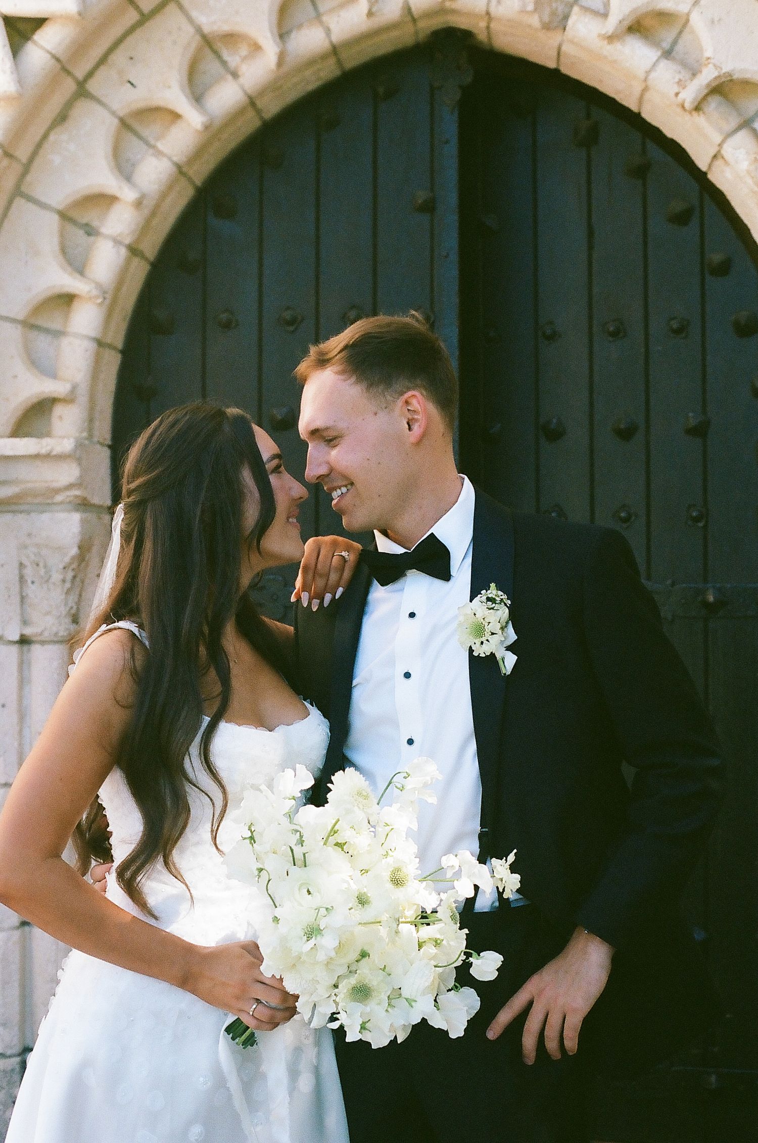 Bride and groom sharing romantic moment before arched wooden church door, white bouquet, black tuxedo.