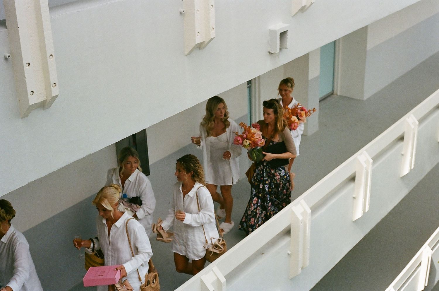 Wedding party members walking down white stairs carrying coral bouquets in casual summer attire.