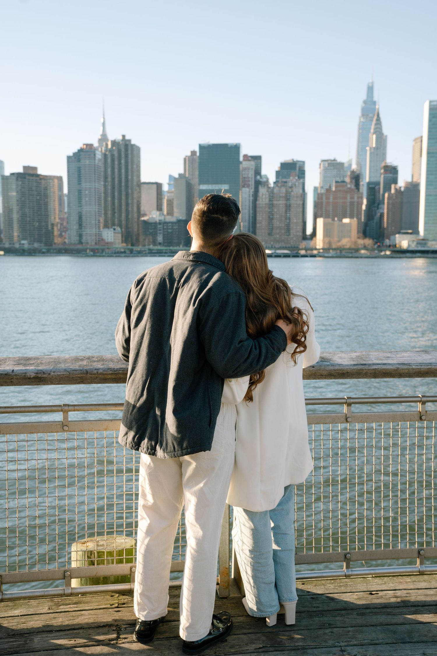 A couple admires the Manhattan skyline while standing on a waterfront boardwalk during daytime.