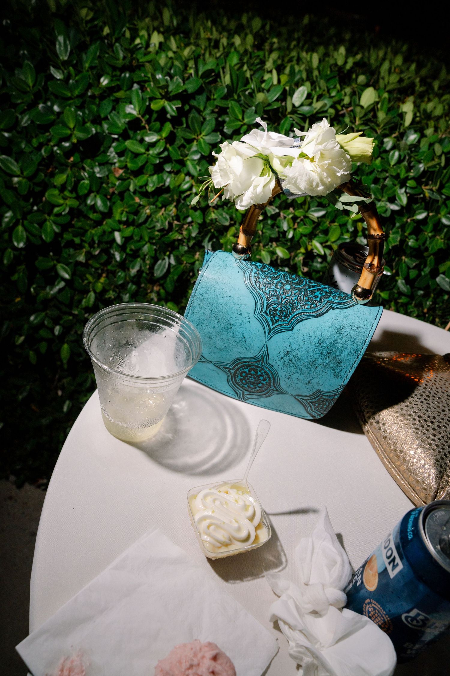 A turquoise handbag and white roses on a table with refreshments against a leafy green hedge background.