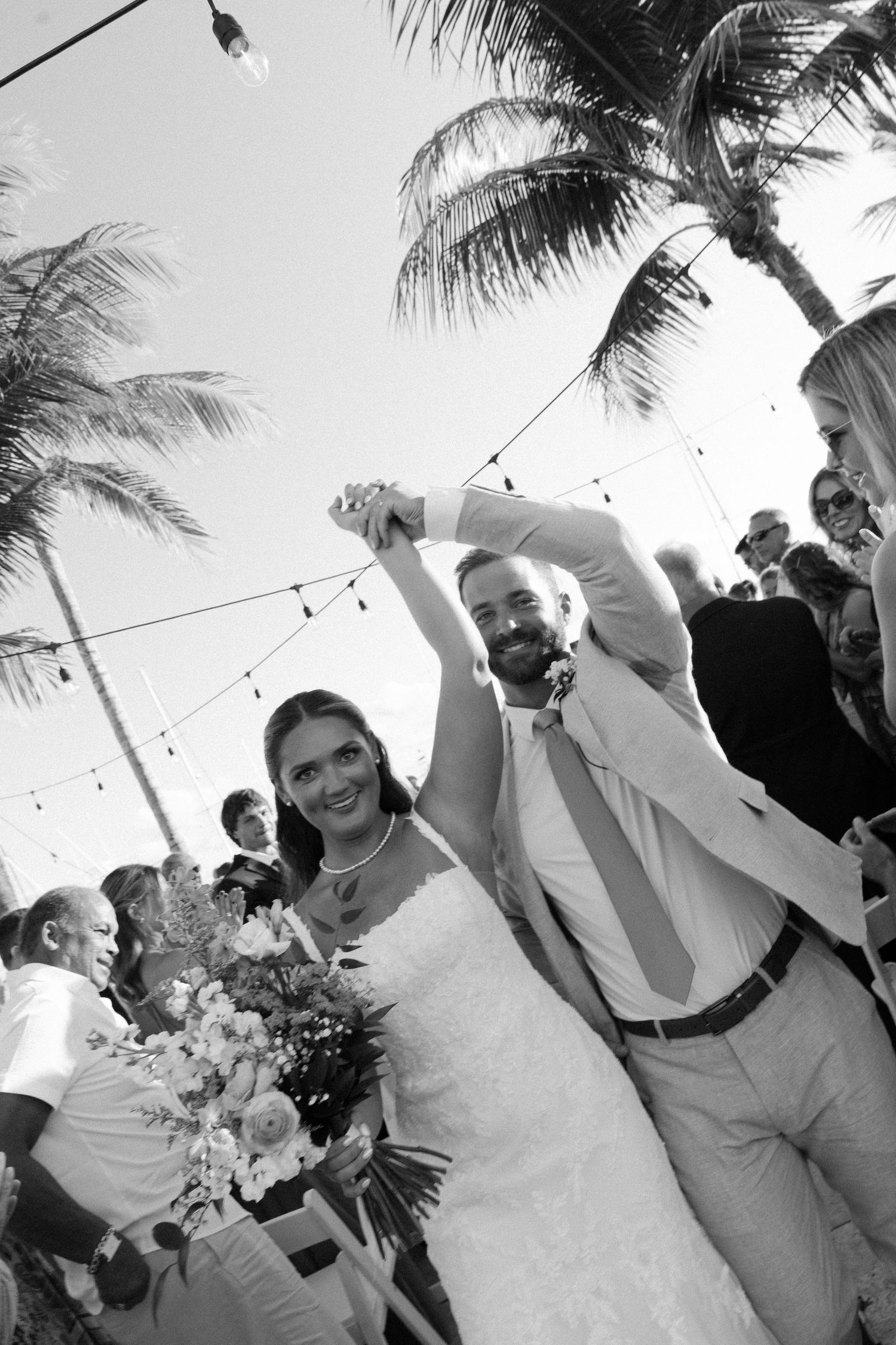 A joyful wedding celebration on a beach with palm trees and string lights as the newlyweds walk down the aisle together.