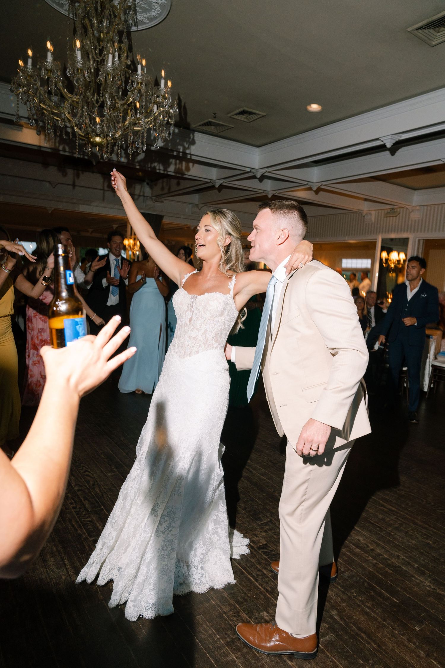 Bride in lace gown and groom in tan suit dancing joyfully at elegant wedding reception under chandelier.