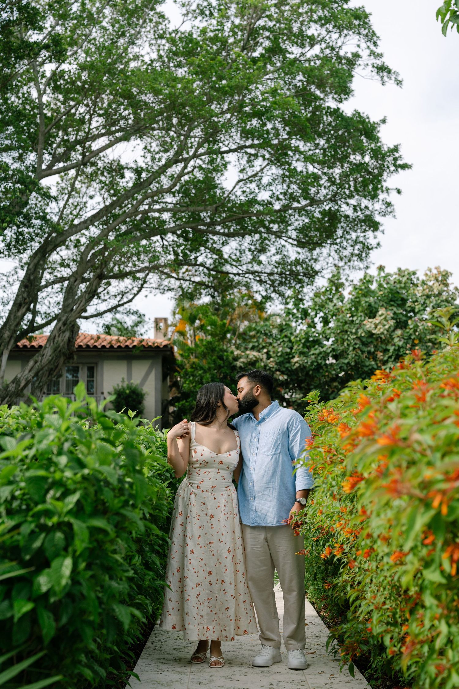 Couple sharing a romantic moment on a garden path surrounded by lush greenery and orange flowers.
