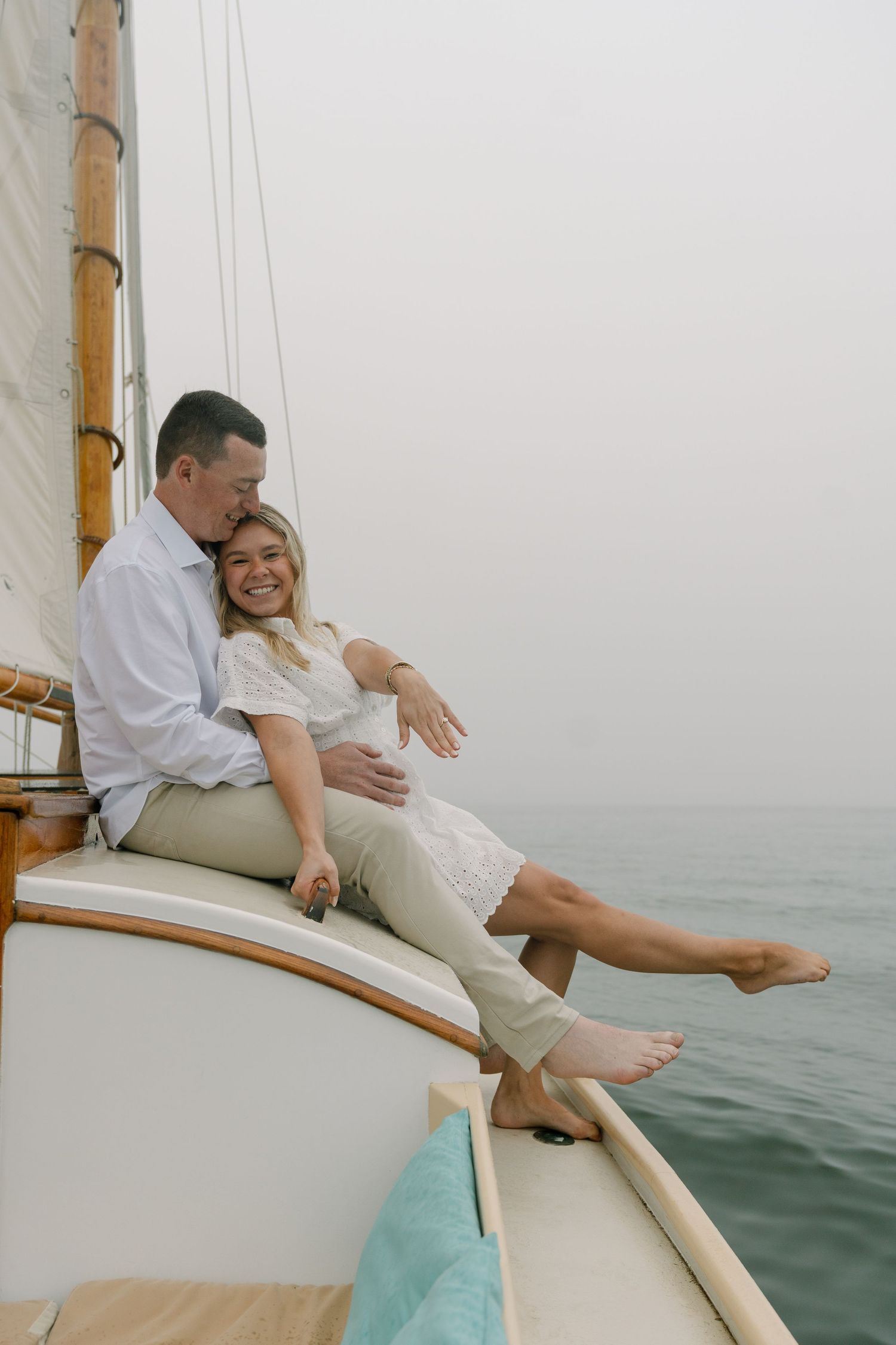 A smiling couple relaxes together on the bow of a sailboat on a calm misty ocean day.