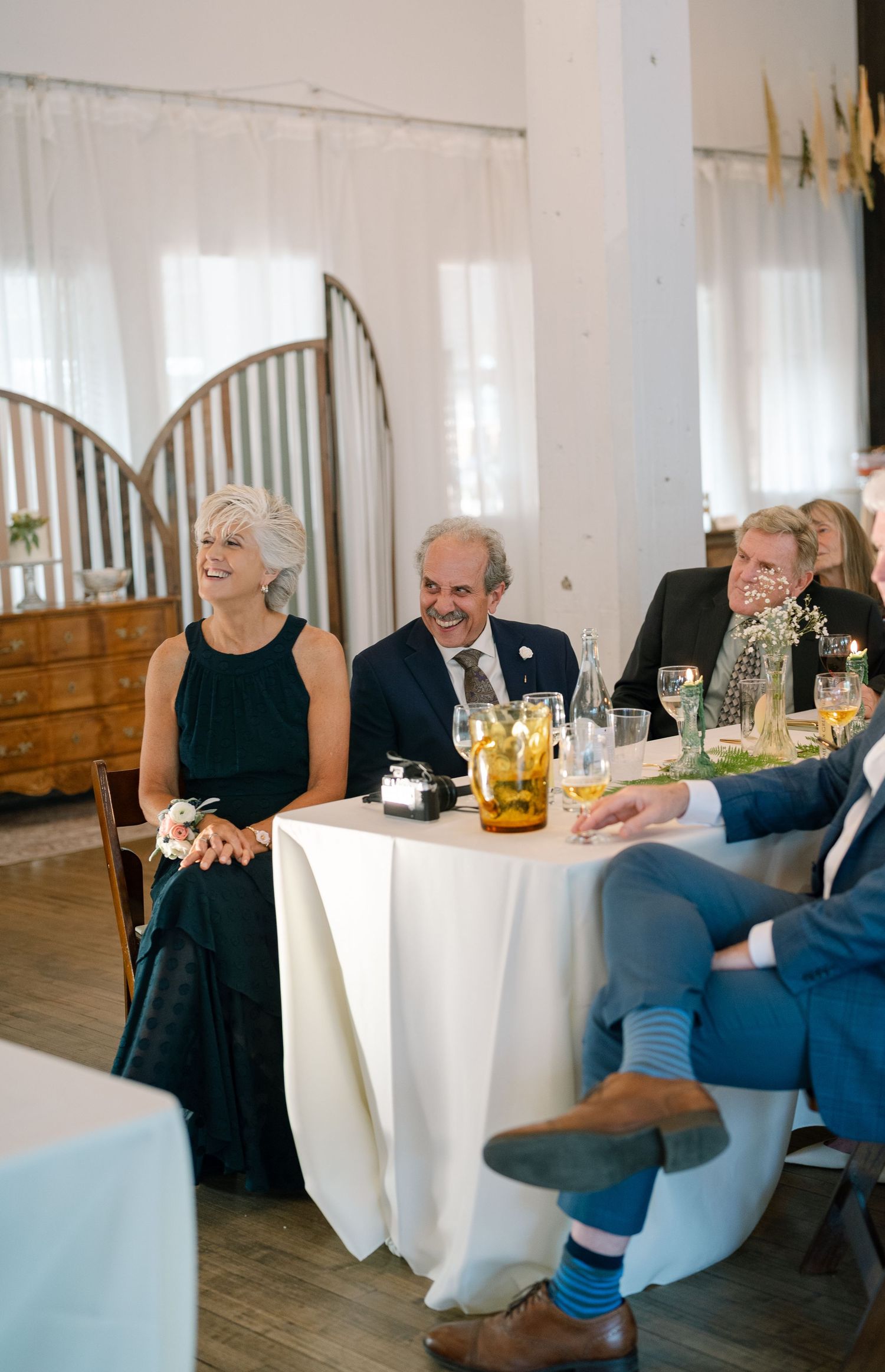 Wedding reception guests enjoying dinner at tables with white linens in bright indoor venue.