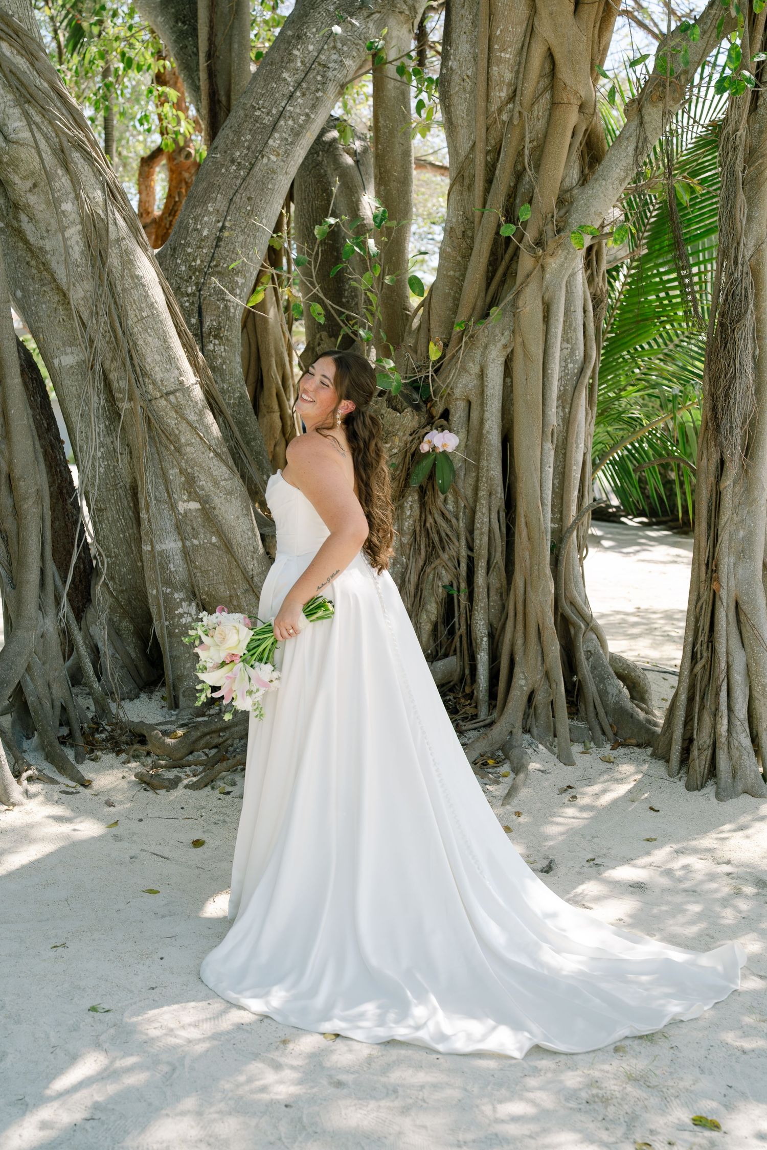 A bride in a white wedding dress stands among the roots of a large tree in a lush, tropical setting.