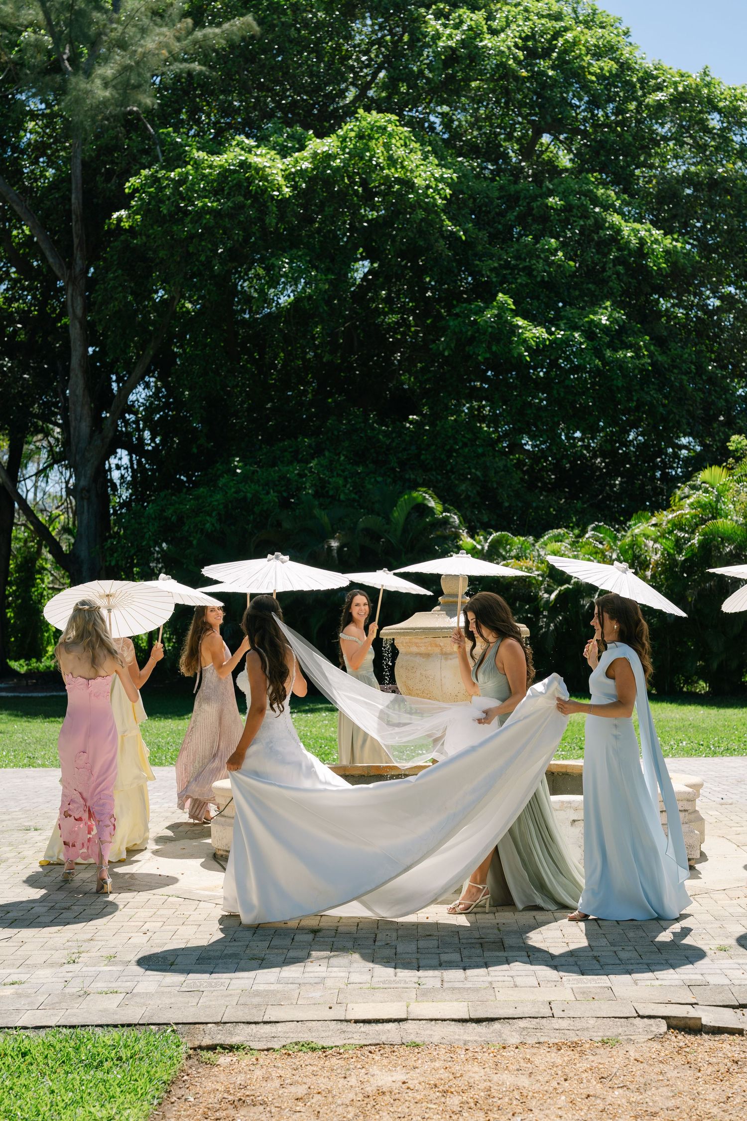 Bridesmaids in pastel gowns hold white parasols while arranging bride's veil outdoors at garden wedding.