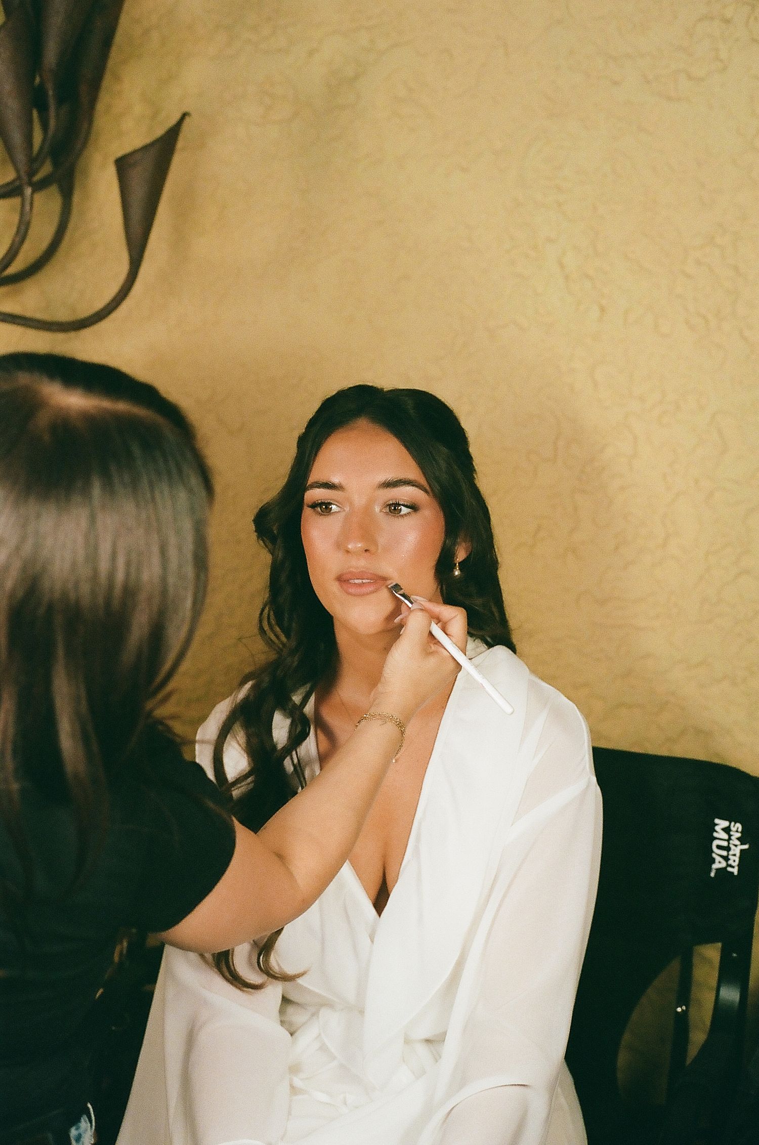 Bride getting makeup applied by artist with brush, wearing white robe, dark wavy hair, preparing for wedding day.