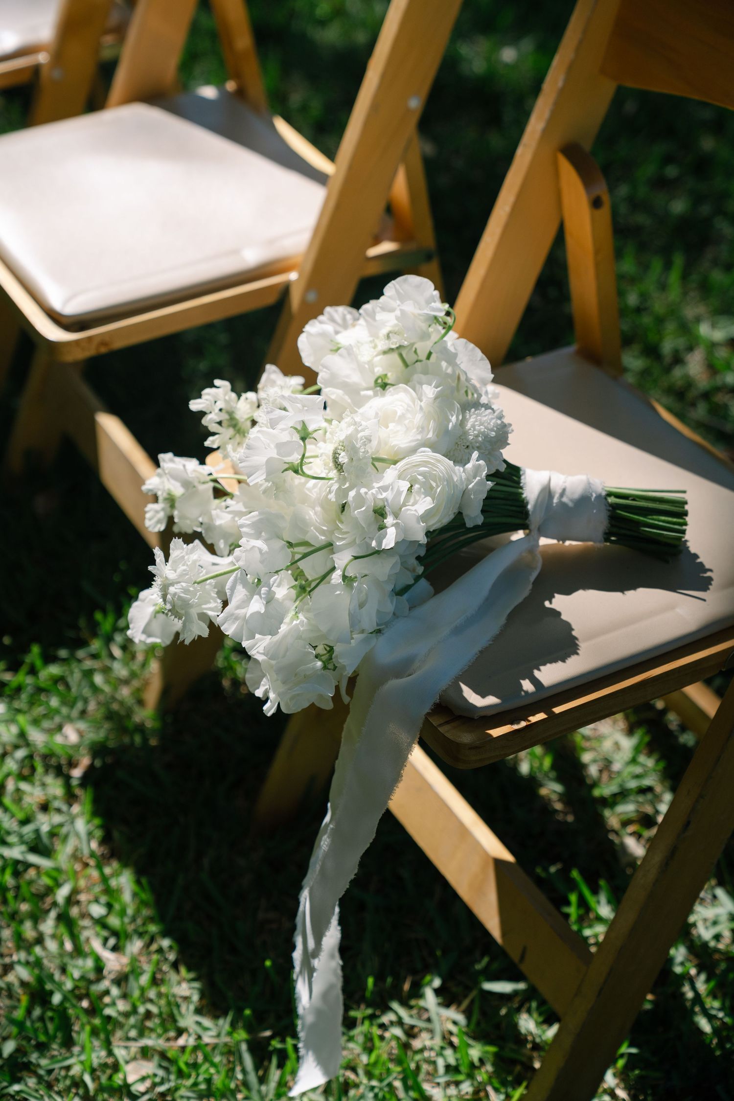 Elegant white wedding bouquet with ribbon rests on wooden folding chair on green grass at outdoor ceremony.