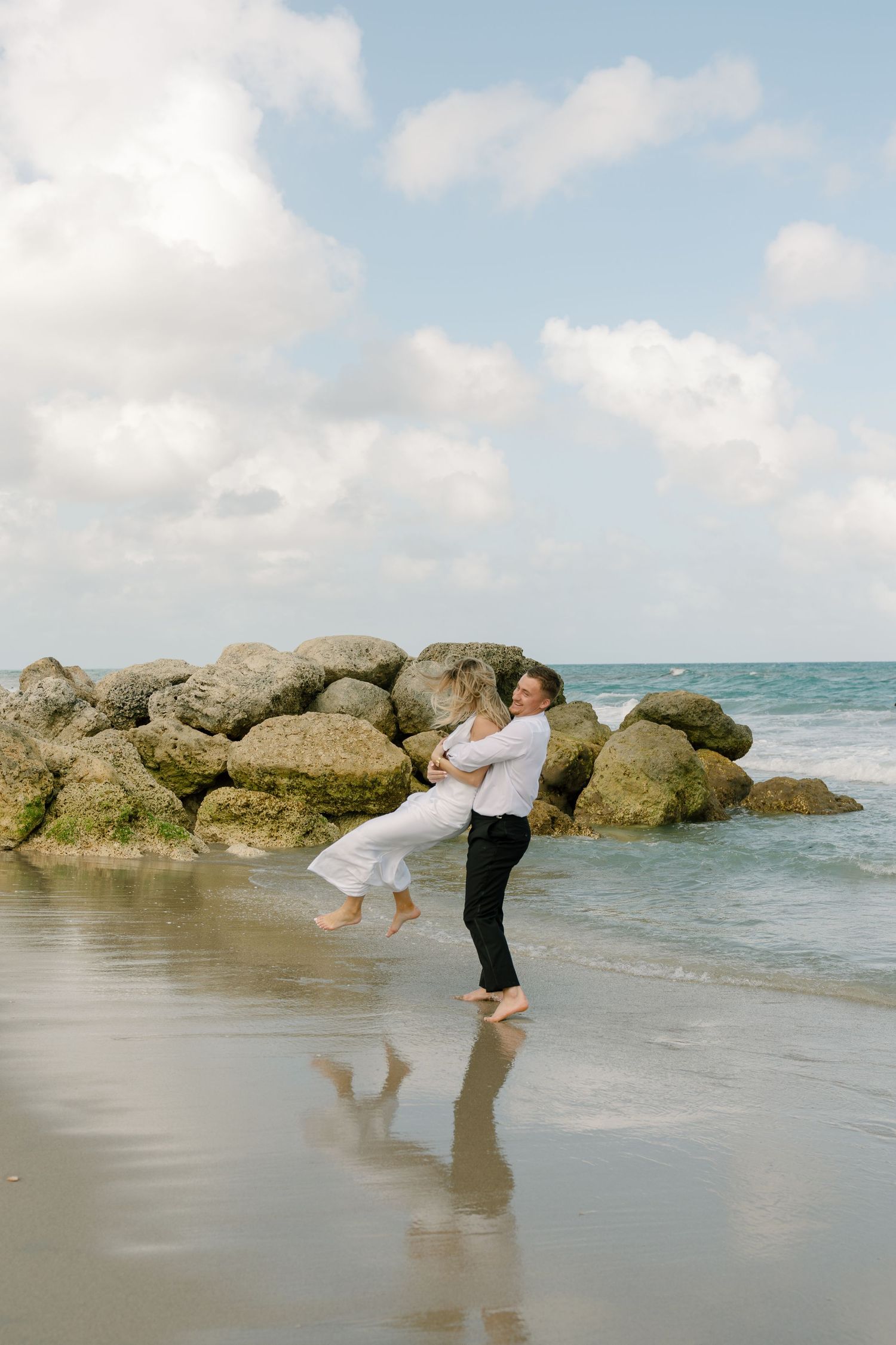 Couple embracing on a sandy beach shore with rocky coastline and ocean waves under a cloudy sky.