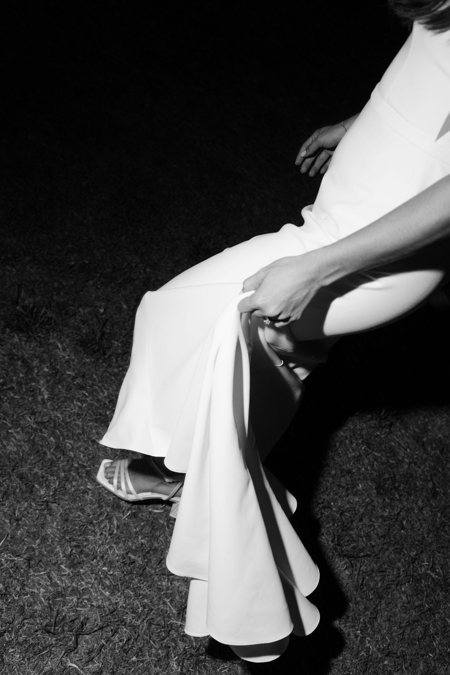 Black and white artistic photo of a bride in a white gown seated outdoors at night showing her heels.