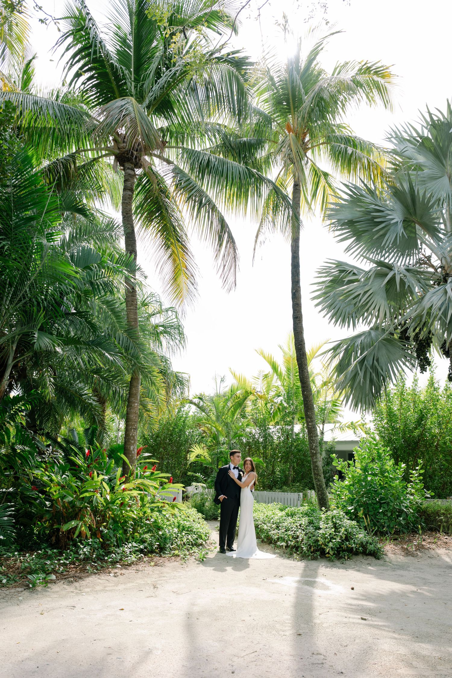 Bride and groom portrait under tall tropical palm trees in lush garden wedding setting.