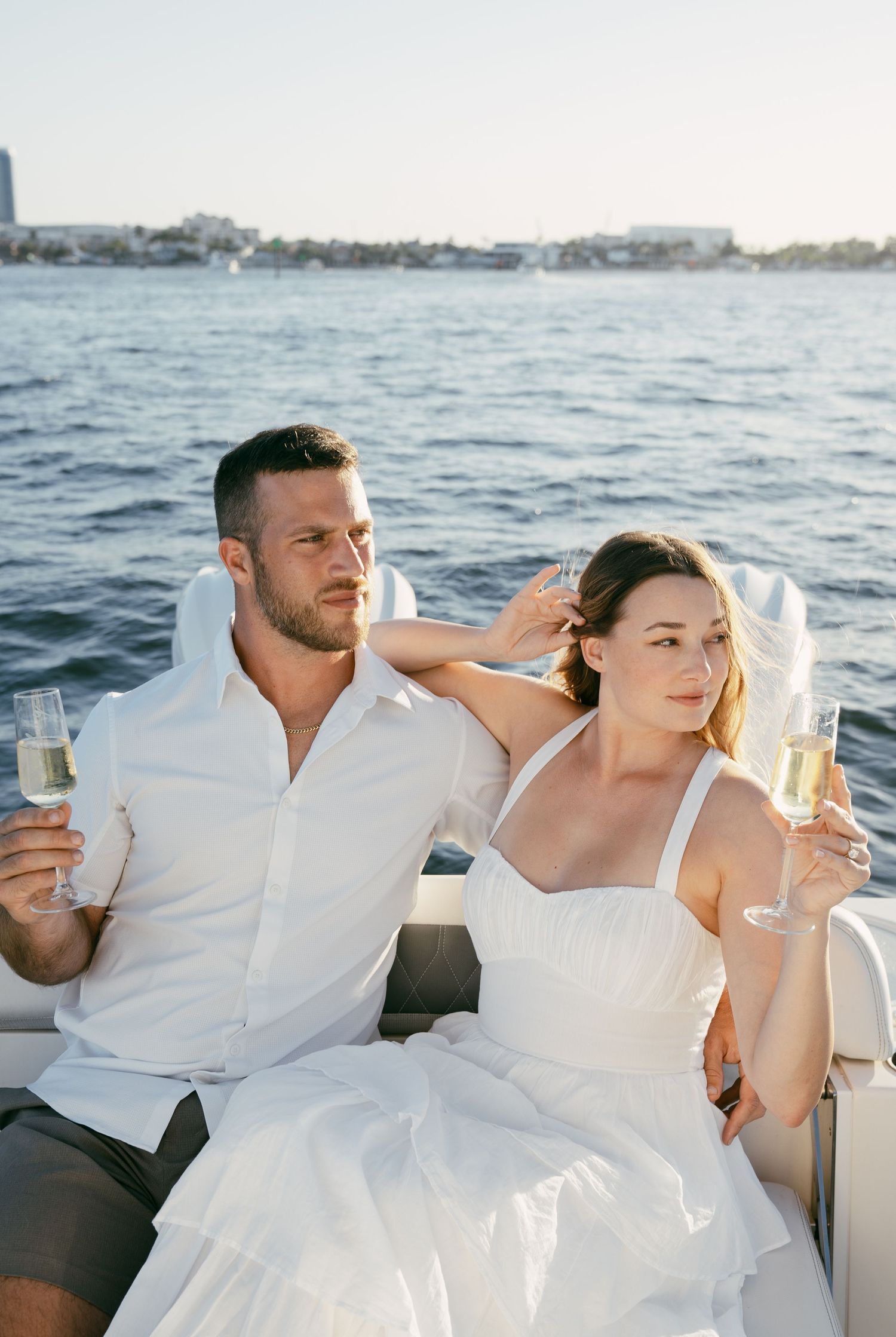 A couple in wedding attire enjoy champagne while sailing on the ocean during sunset.